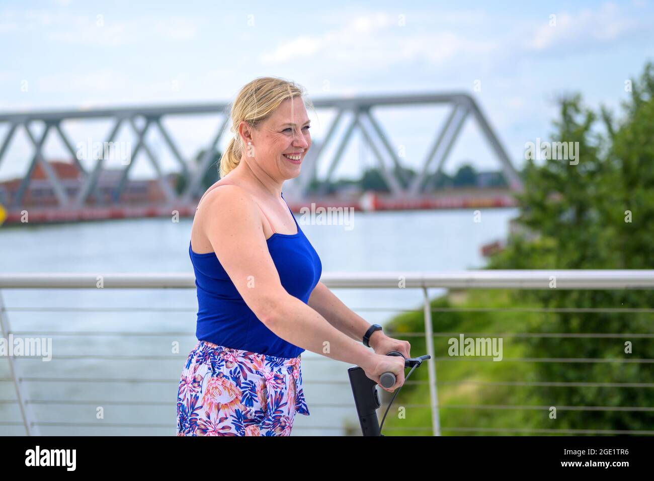 Happy vivacious middle-aged blond woman on an electric scooter crossing a bridge over a river on a hot summer day in an urban mobility or transportati Stock Photo