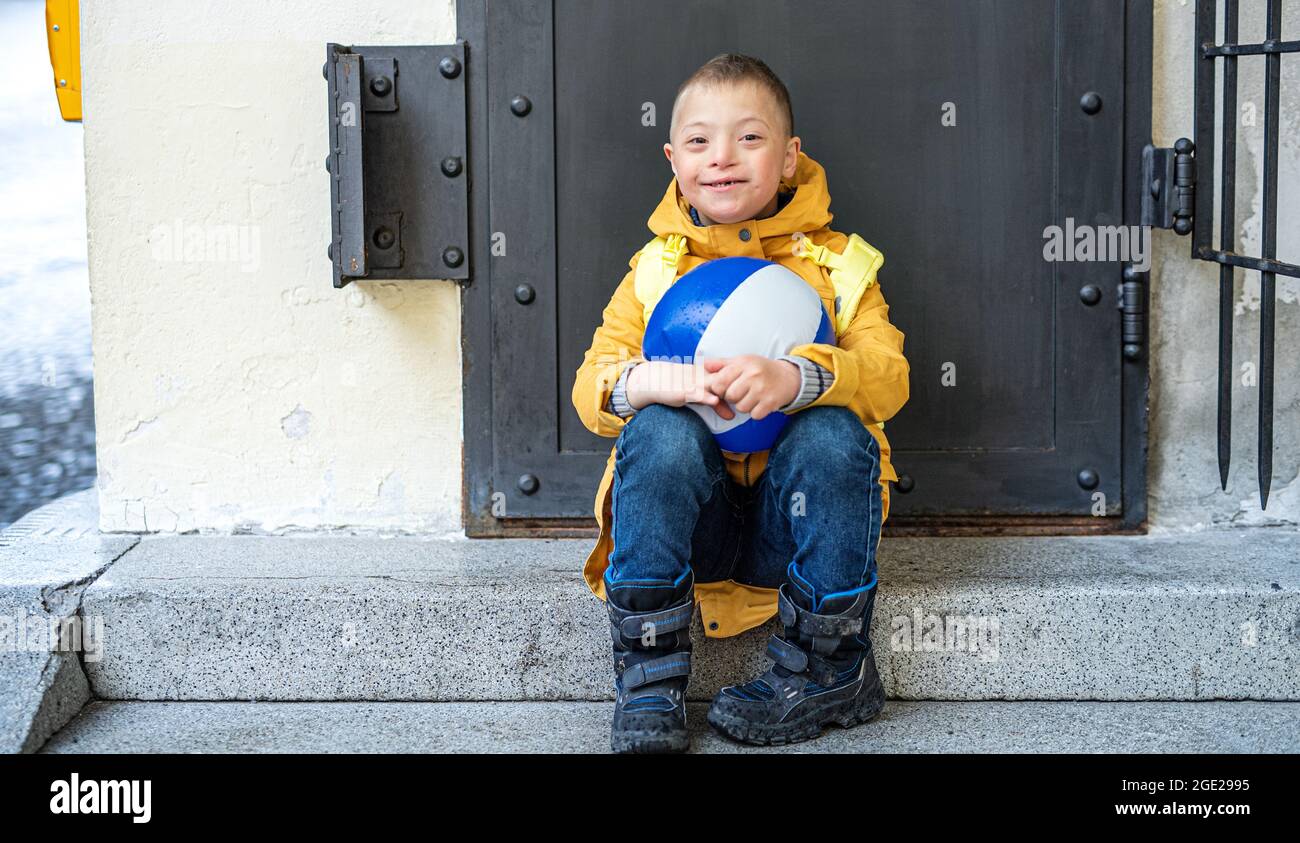 Happy down syndrome boy with ball outdoors sitting in front of door, looking at camera. Stock Photo