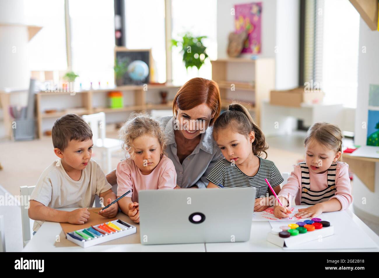 Group of small nursery school children with teacher indoors in classroom, using laptop. Stock Photo