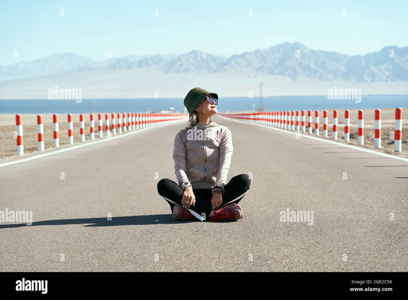 asian woman tourist sitting in the middle of an empty open road looking at view with lake and rolling mountains in background, leg crossed. Stock Photo