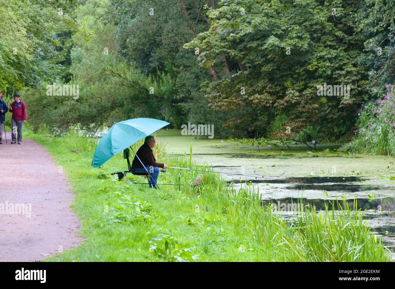 Walker on a path next to a senior man under a green umbrella fishing in a watercourse near Ashton under Lyne, Greater Manchester, UK Stock Photo
