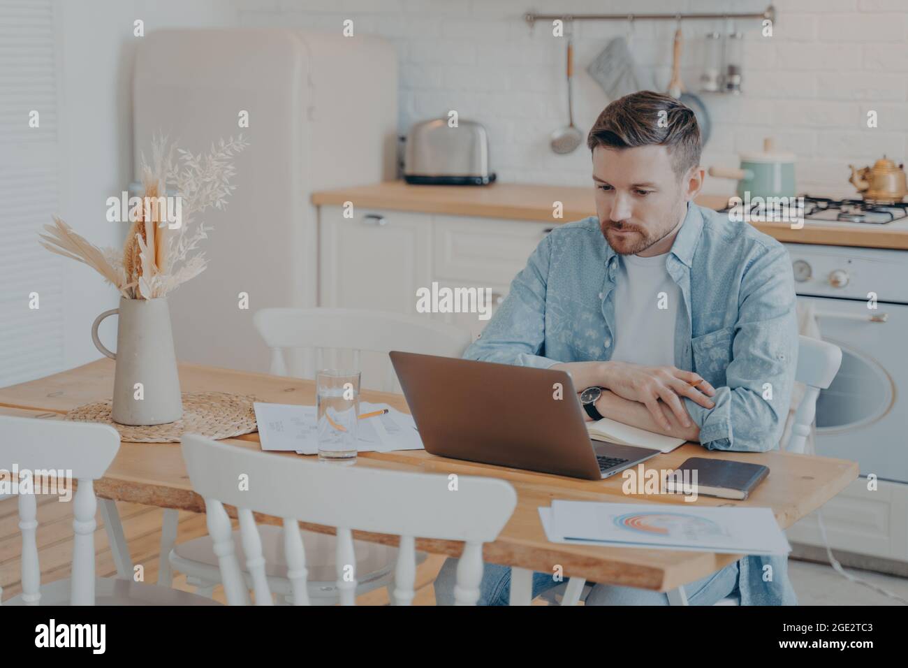 Focused young freelancer working on laptop while sitting by kitchen table Stock Photo