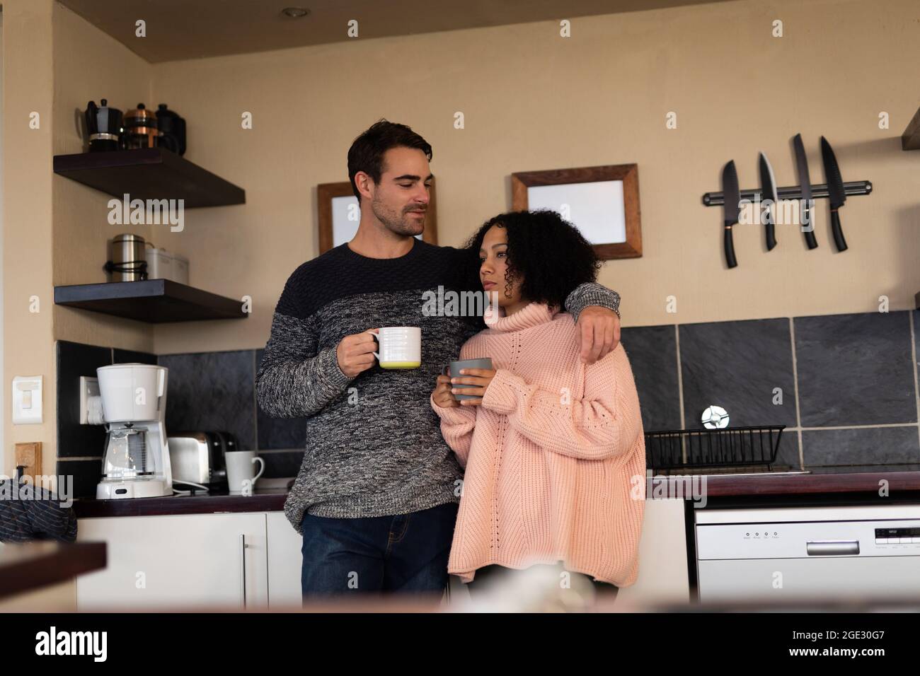 Happy diverse couple in kitchen embracing drinking coffee and smiling Stock Photo