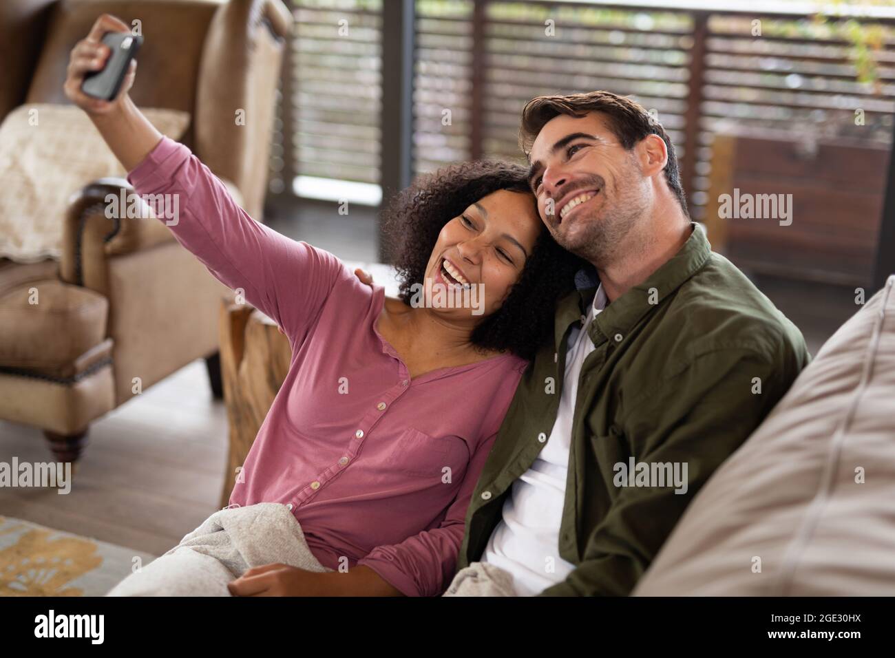 Happy diverse couple sitting on sofa in living room taking selfie and smiling Stock Photo