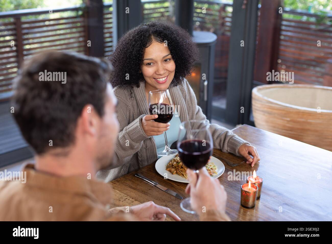 Happy diverse couple in living room sitting at table eating dinner together and drinking wine Stock Photo