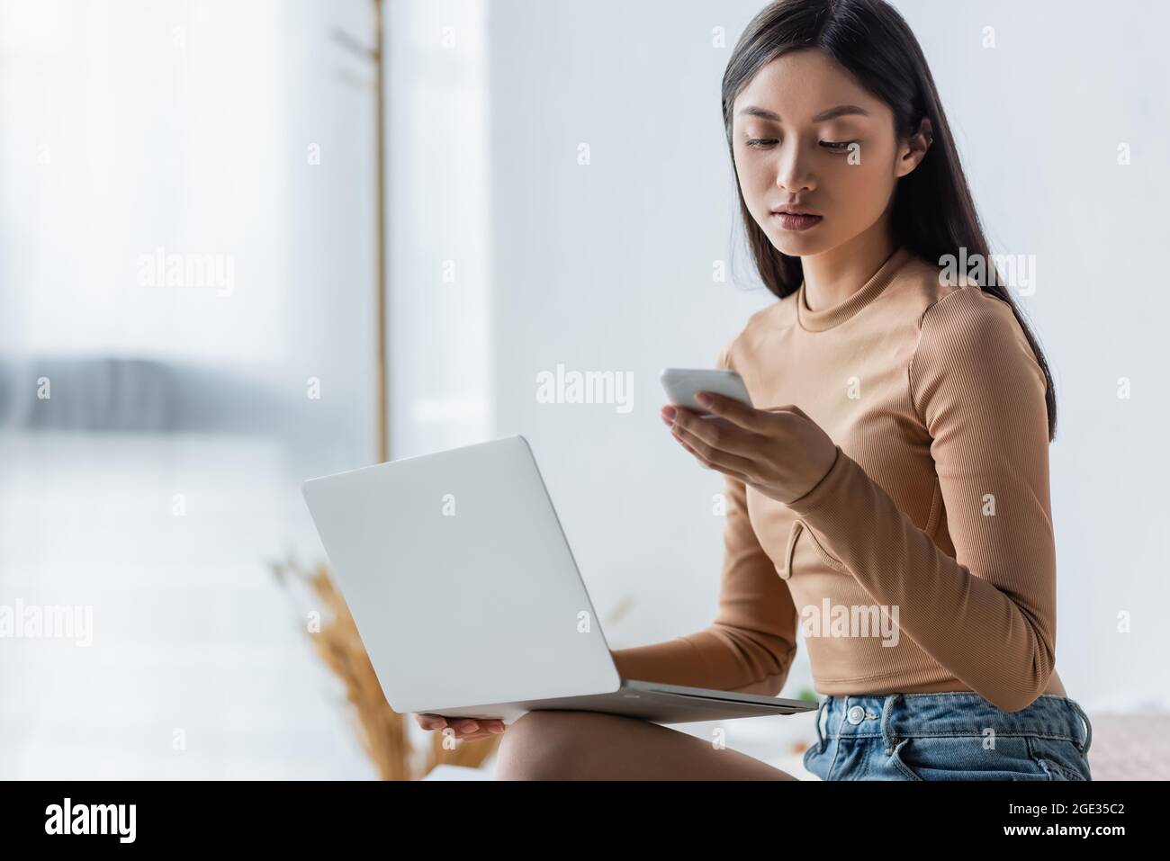 young asian woman looking at mobile phone while working near laptop at ...