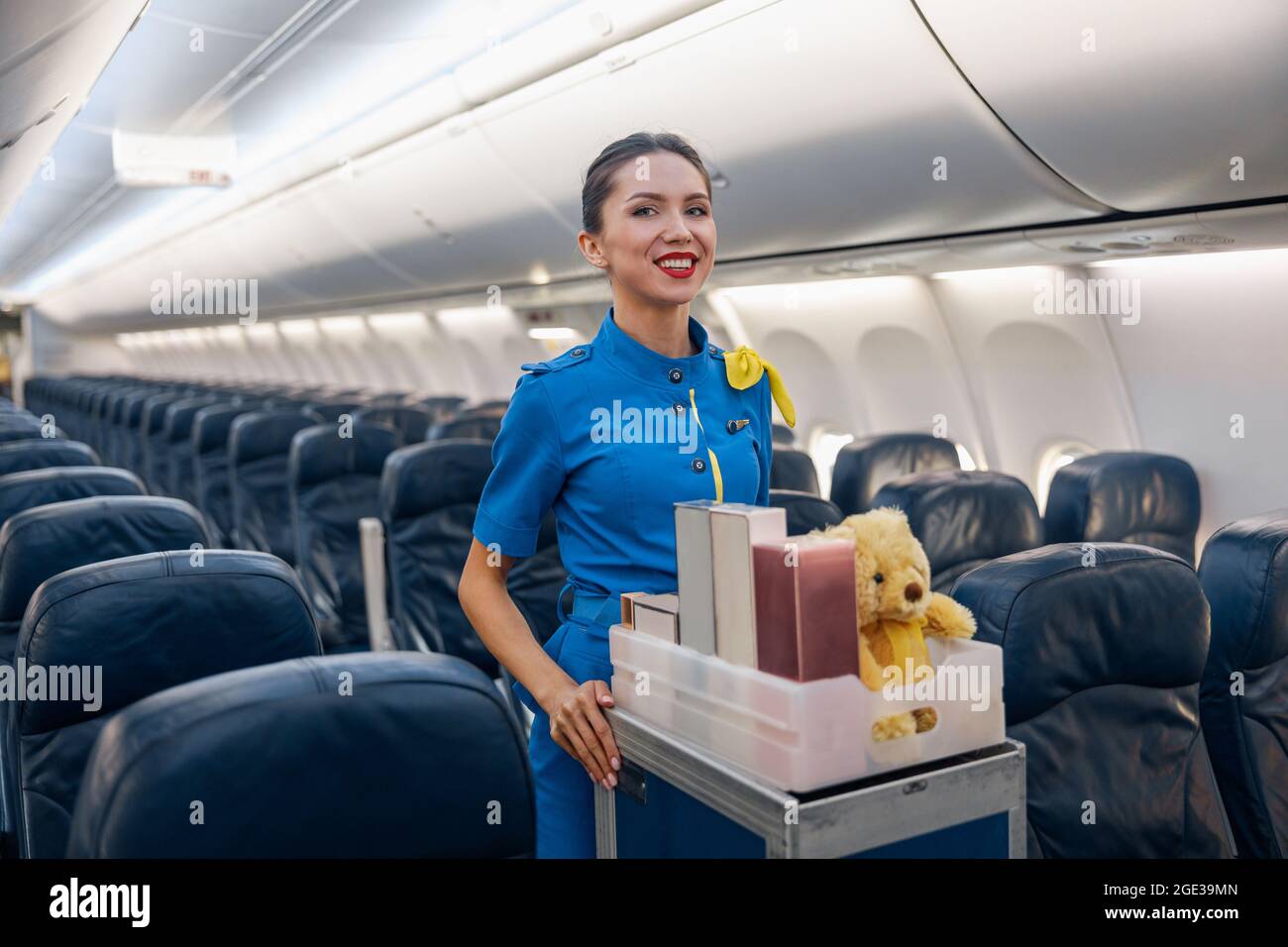 Cheerful female air hostess in bright blue uniform smiling at camera while leading trolley cart with gifts through empty plane aisle Stock Photo