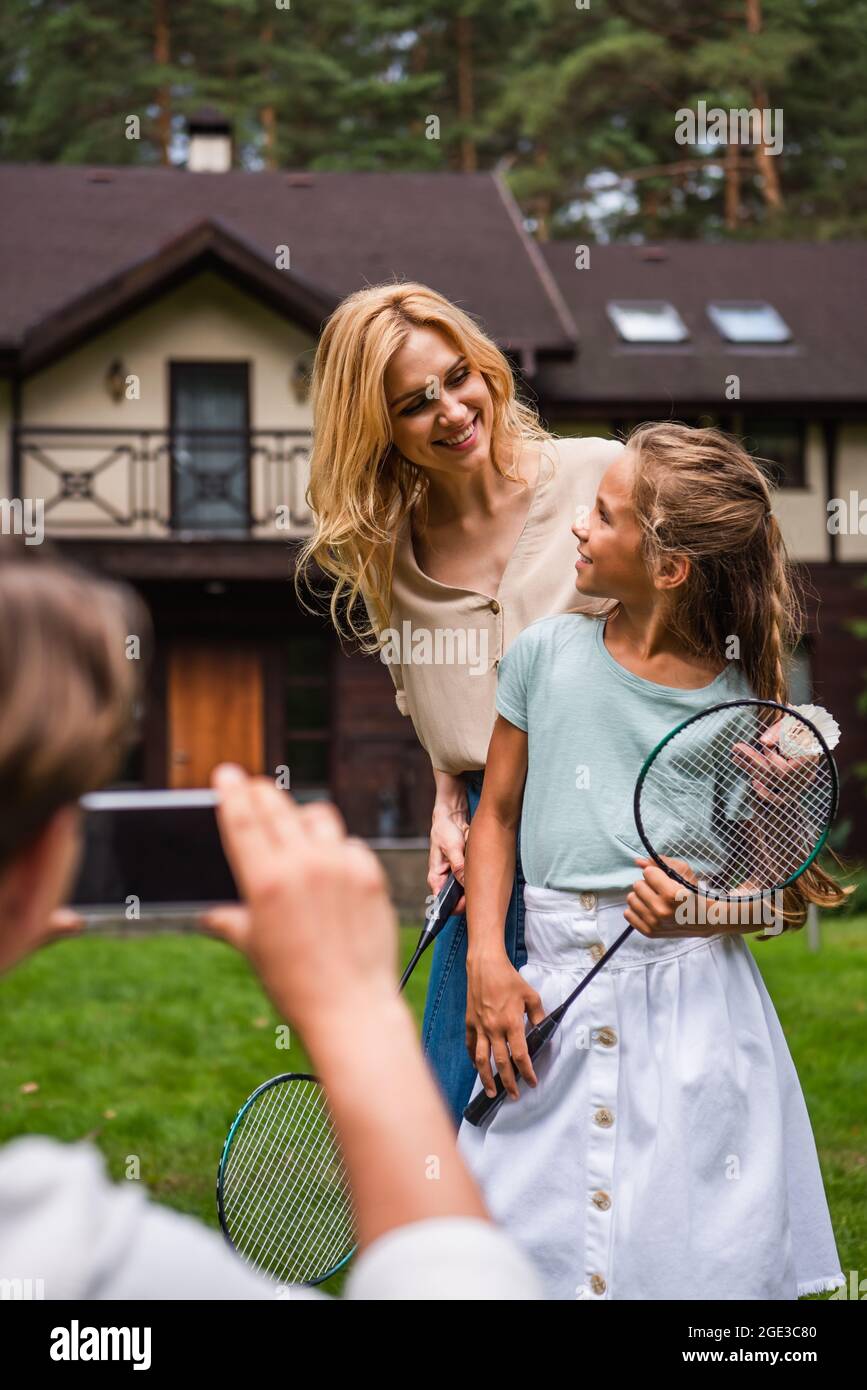 Smiling mother and daughter with badminton rackets standing near blurred father with smartphone Stock Photo