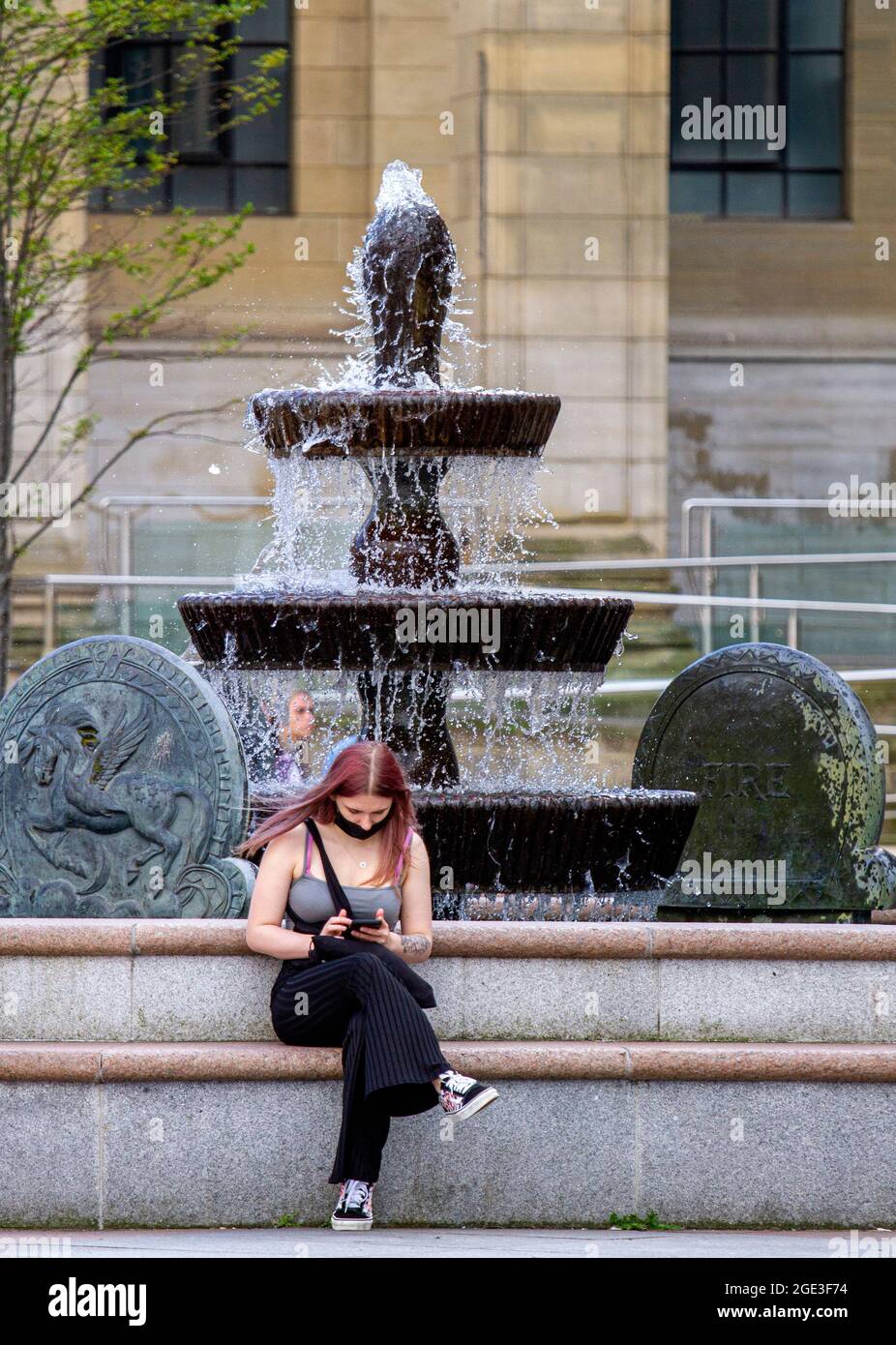 Dundee, Tayside, Scotland, UK. 16th Aug, 2021. UK Weather: A warm sunny day with a slight cool breeze across North East Scotland with temperatures reaching 20°C. A young woman wearing a facemask sitting at the Caird Hall fountains enjoying the warm sunshine whilst text messaging on her mobile phone in Dundee city centre. Credit: Dundee Photographics/Alamy Live News Stock Photo