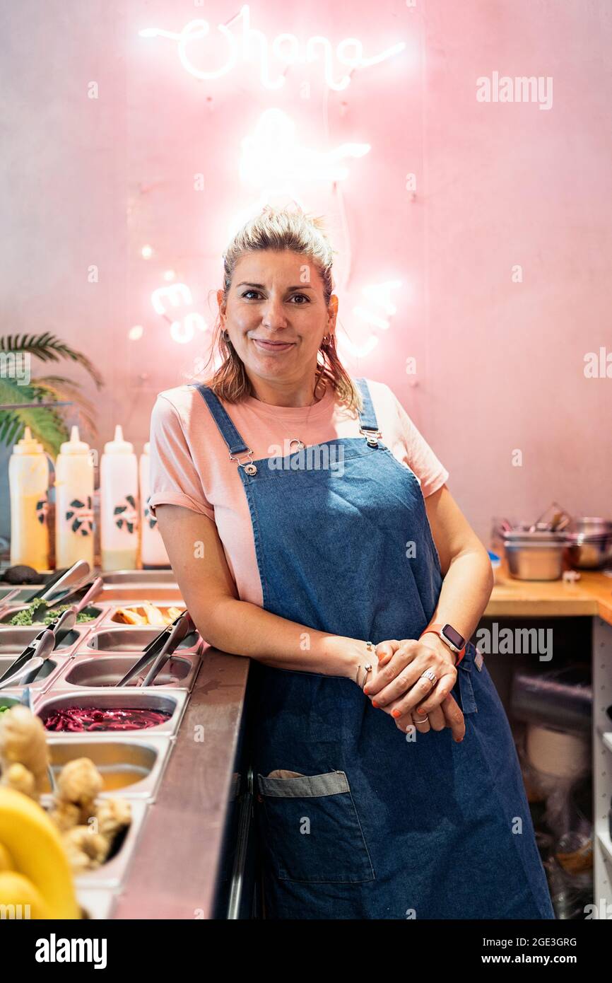 Blonde woman with apron smiling and looking at camera while working in ...