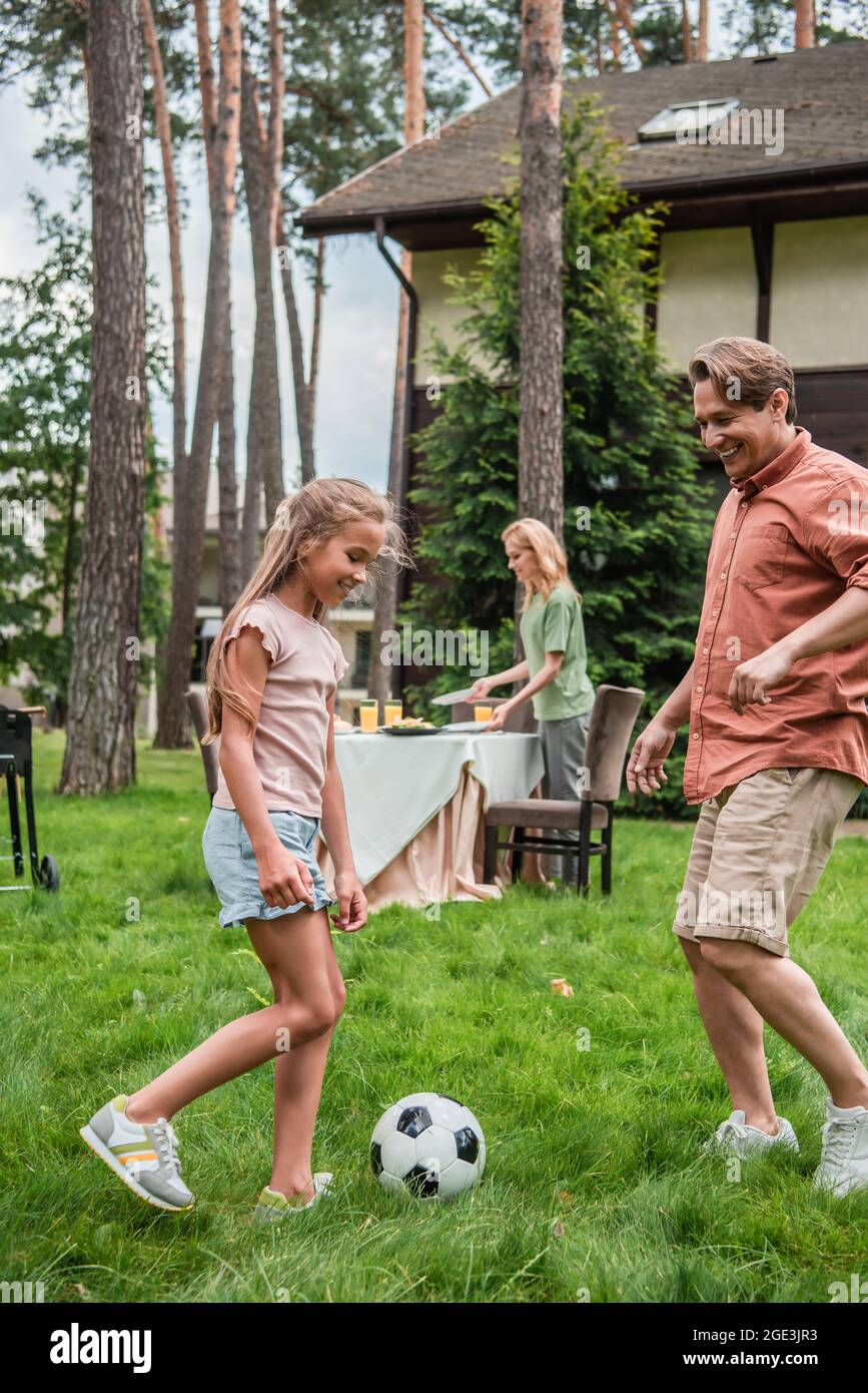 Smiling kid playing football with father near mother and table outdoors Stock Photo