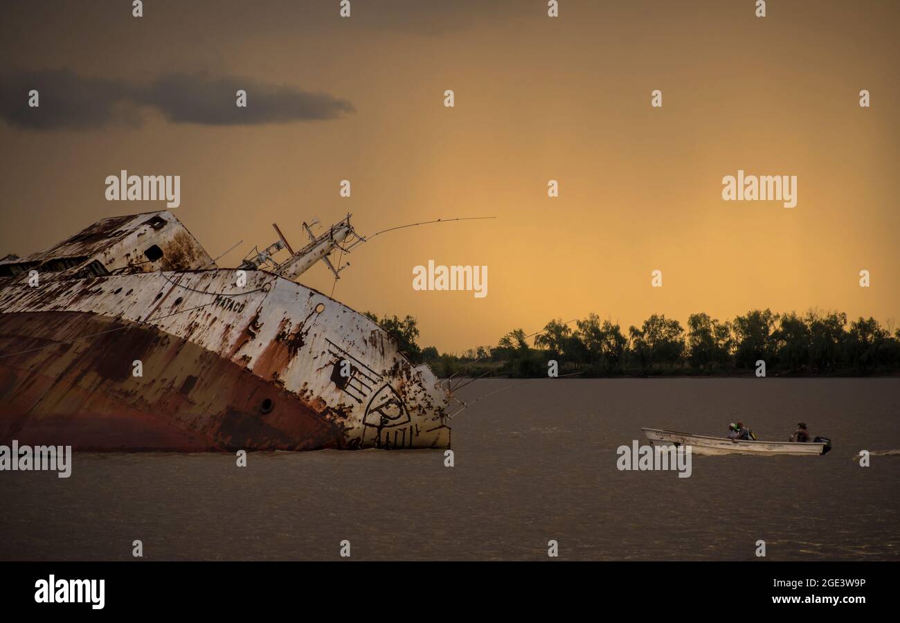 Big sunken ship in the river "Mataco" with a little boat siling next to it with an orange sky at sunset Stock Photo