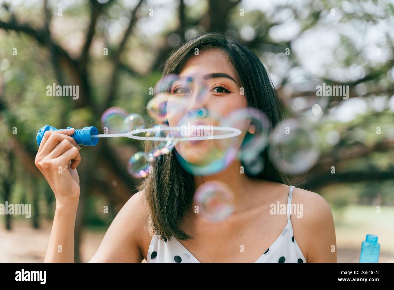 South East Asian ethnic 20s woman blowing air bubbles in the park Young Happy girl wears stylish dress with trees in background. Outdoor summer weekend activity - with copy space Stock Photo