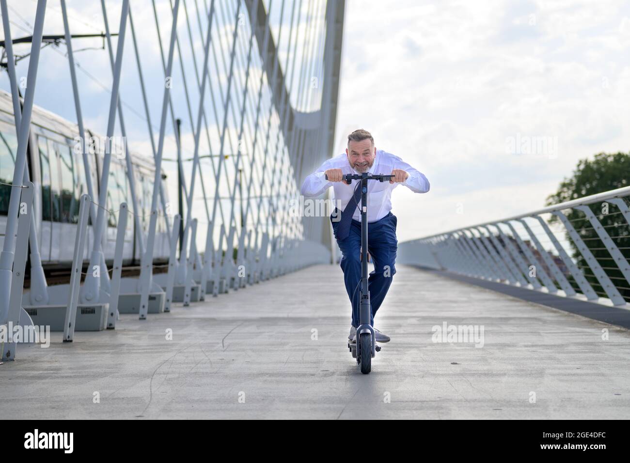 Middle-aged man crouching low over the handlebars with a happy smile as he speeds along on an electric scooter crossing a bridge towards the camera Stock Photo