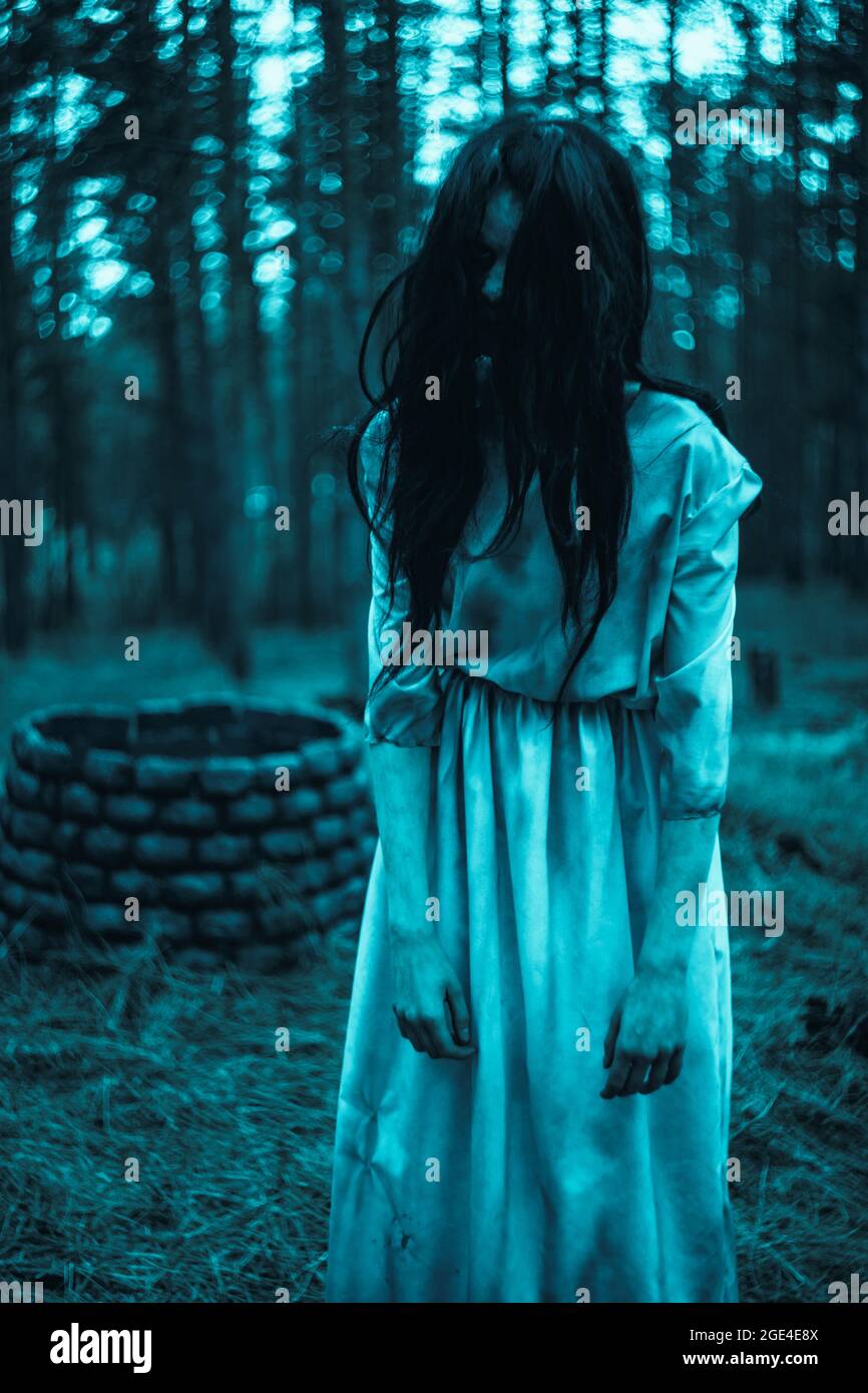 Girl with long black hair in image of scary ghost zombie stands among dark  forest against background of stone well Stock Photo - Alamy