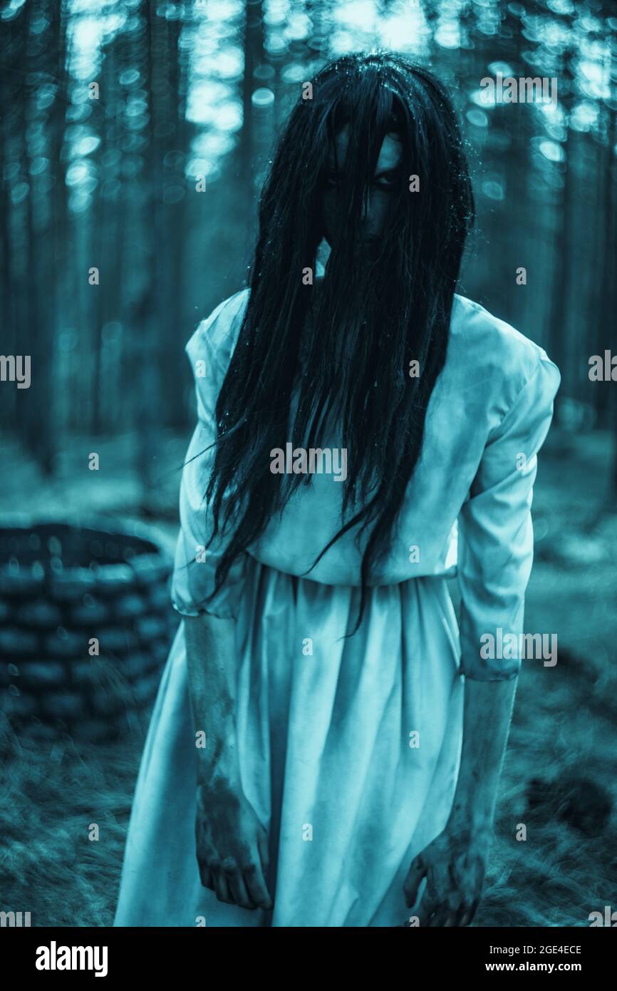 Girl with long black hair in image of scary ghost zombie stands among dark  forest against background of stone well Stock Photo - Alamy