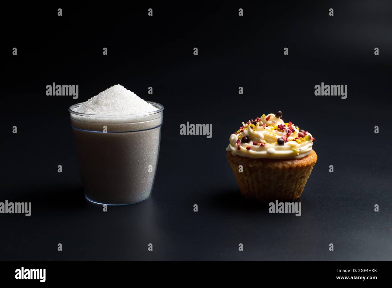 Cupcake and cup of sugar close up, on black background. Food, which contains a large amount of sugar, harmful to health. Stock Photo