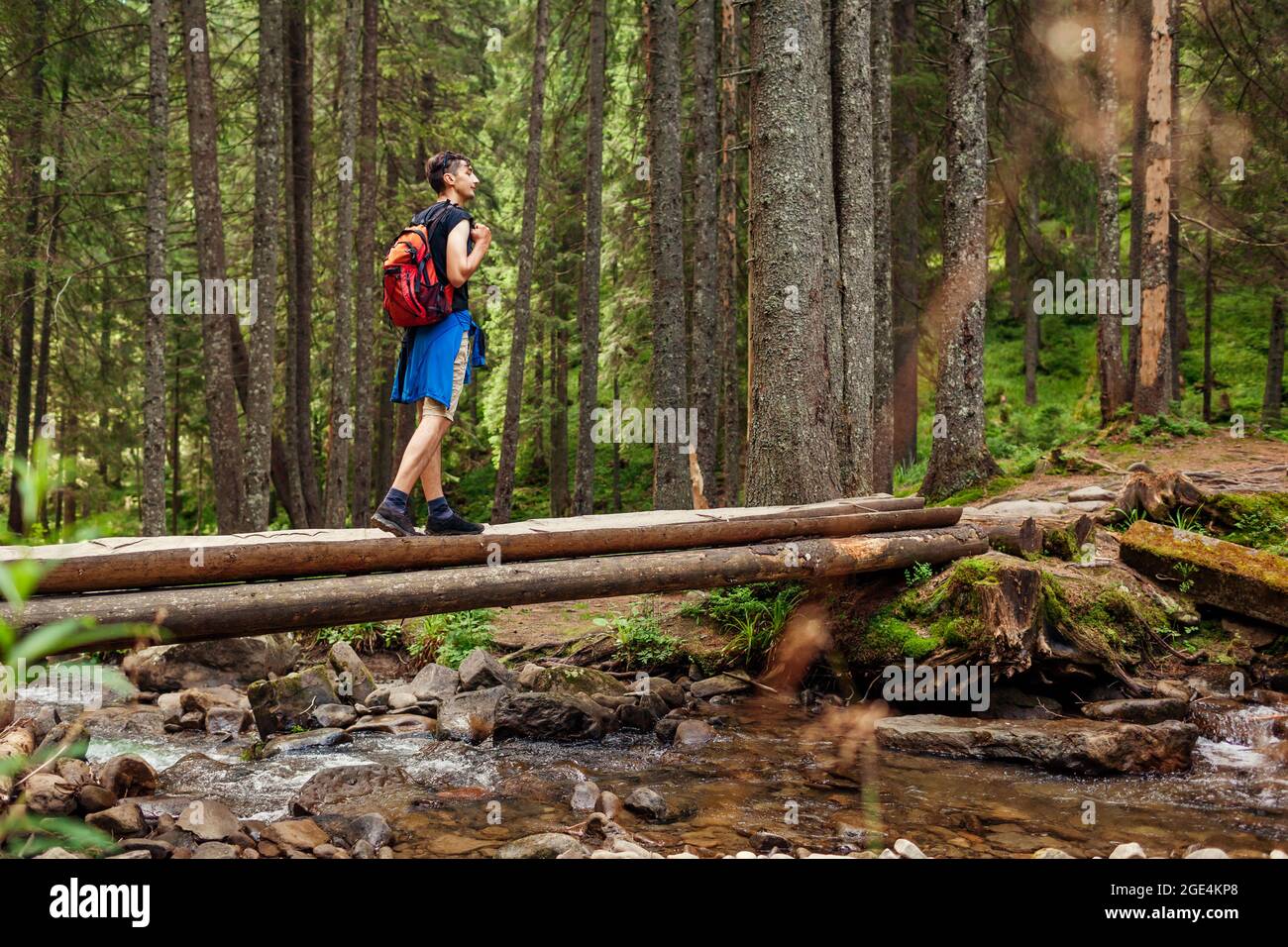 Traveler hiker with backpack crossing mountain river in Carpathian forest walking on wooden bridge. Man tourist enjoying landscape. Trip to summer Ukr Stock Photo