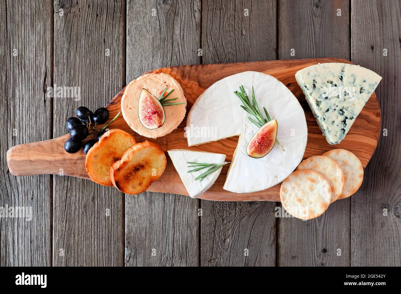 Cheese board with a selection of cheeses, crackers, figs and grapes on wooden serving board. Above view on a rustic wood background. Stock Photo
