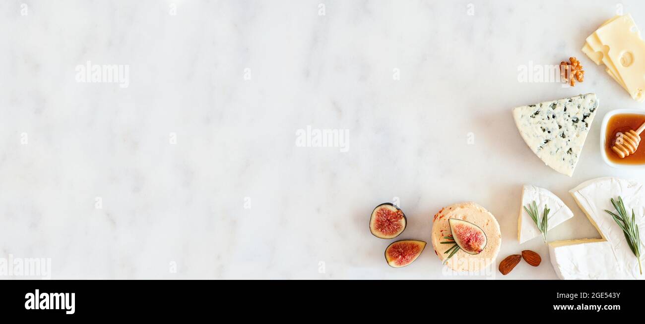 Corner border of a selection of cheeses, figs, nuts and honey. Above view banner on a white marble background with copy space. Stock Photo