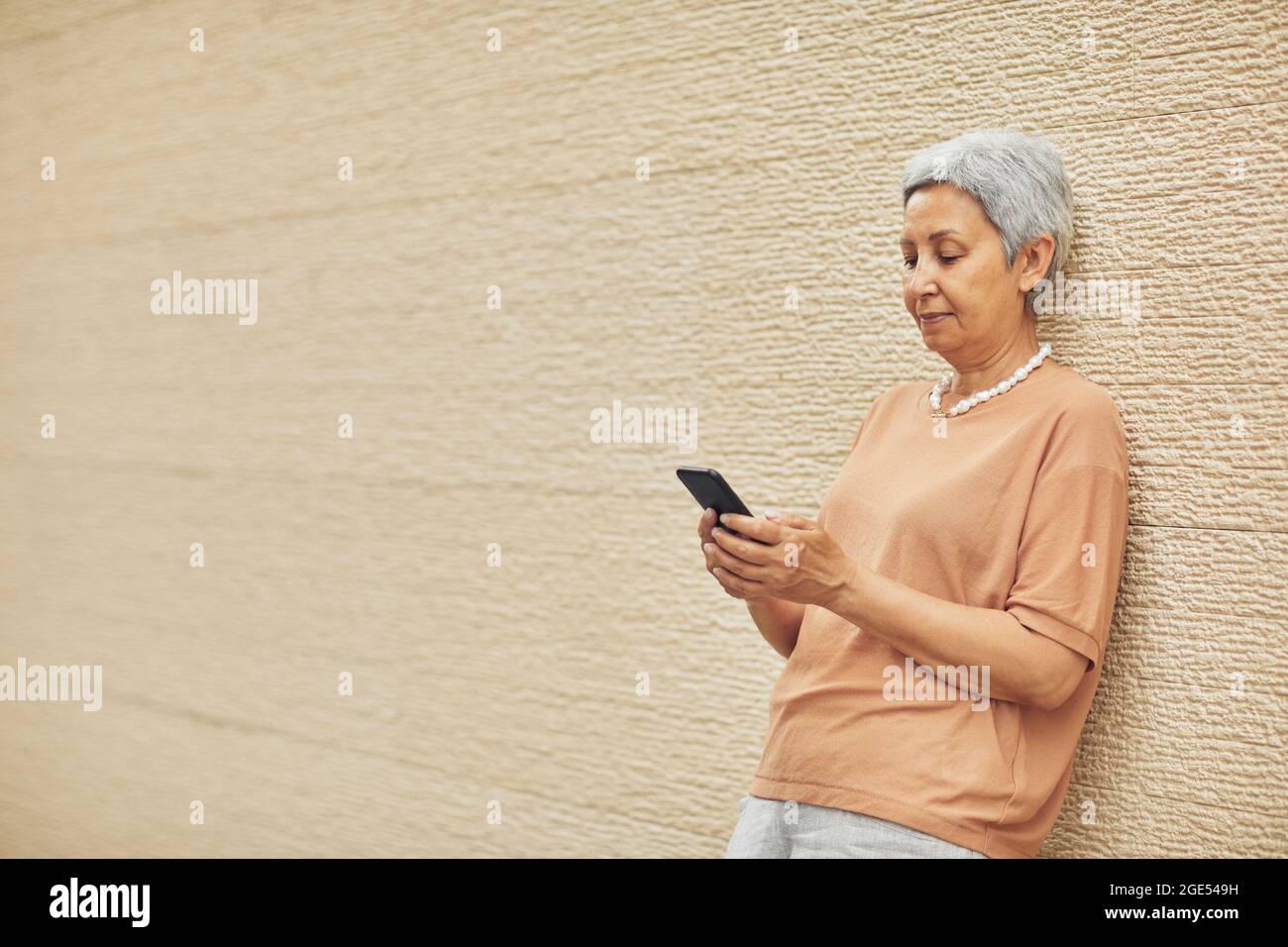 Minimal side view portrait of modern mature woman using smartphone while standing against yellow wall outdoors, copy space Stock Photo