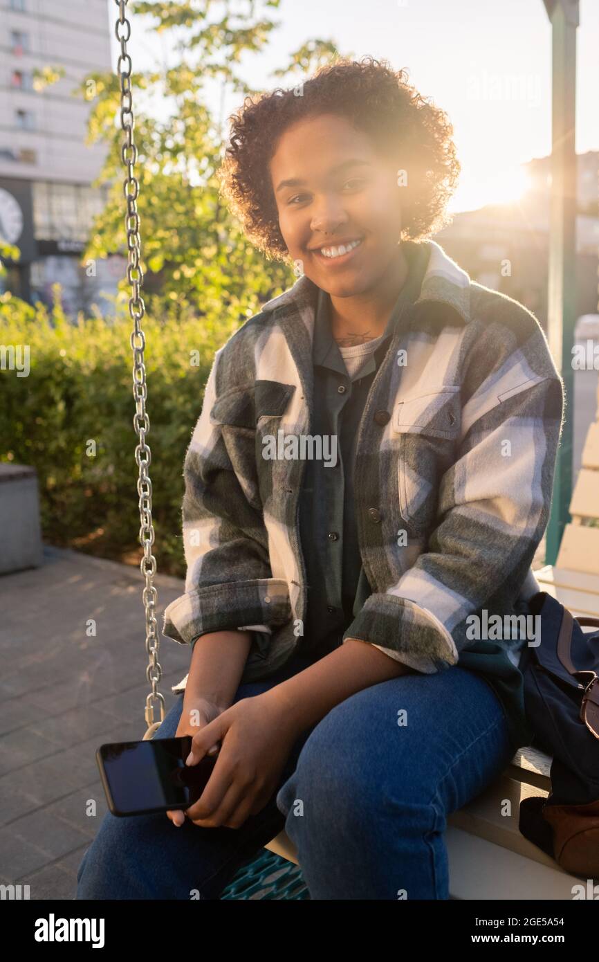 Cheerful African teenager with smartphone sitting on swing and looking at you with smile Stock Photo