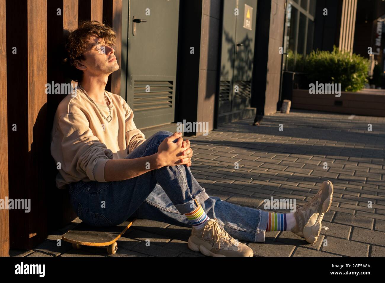 Young relaxed man in casualwear sitting on skateboard at sundown Stock Photo