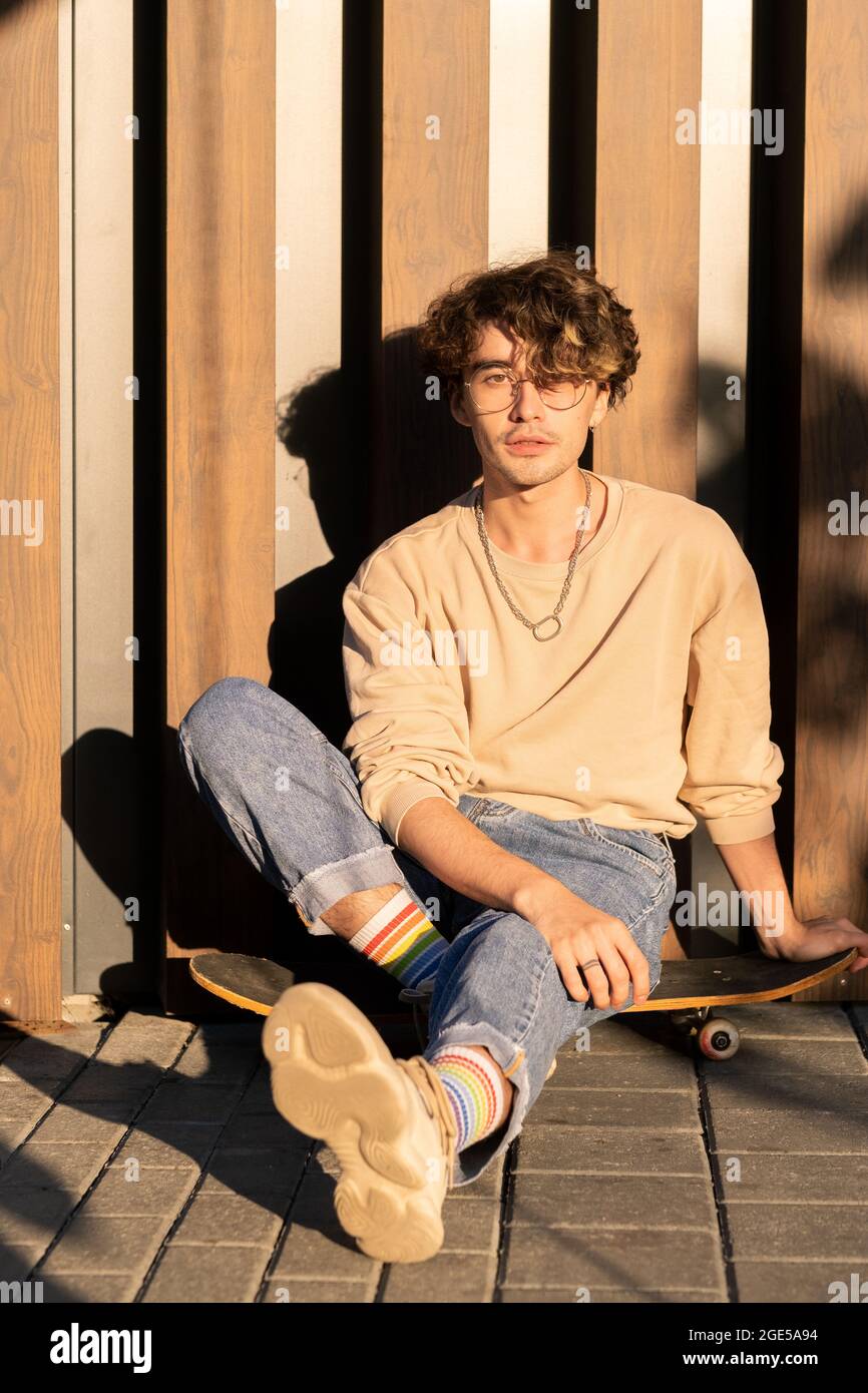 Handsome guy in jeans and sweatshirt sitting on skateboard by wall Stock Photo