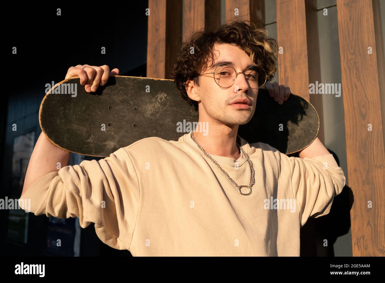 Teenage guy holding skateboard behind his neck while standing by building exterior Stock Photo