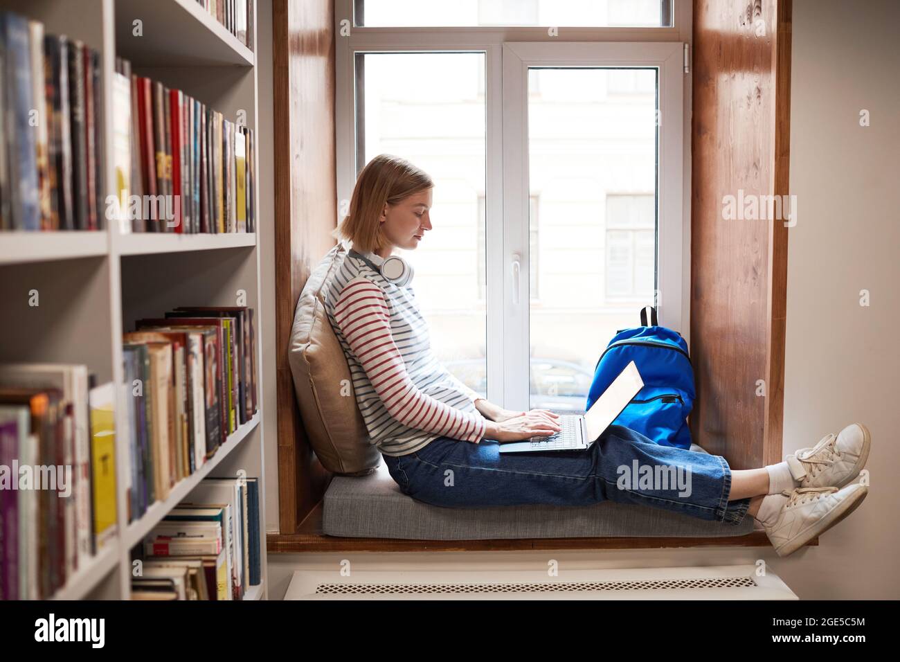 Full length side view portrait of young woman relaxing by window at reading lounge in library, copy space Stock Photo