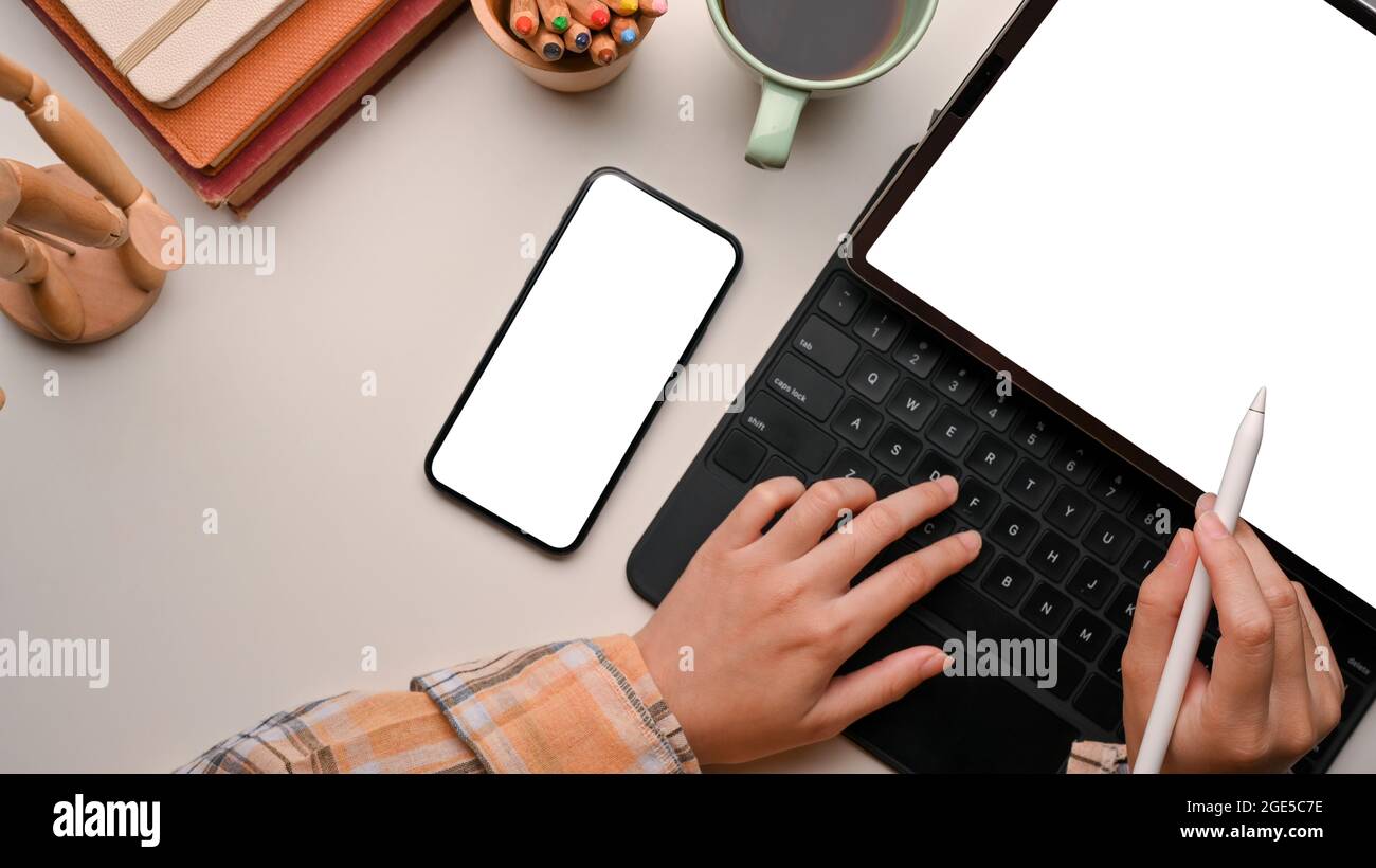 Minimal hipster feminine workspace, female typing on tablet, tablet blank screen mockup, smartphone mockup, coffee, colour pencils, books, figure on n Stock Photo