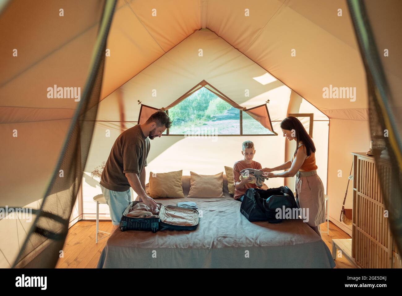Mother passing stack of clothes to her teenage son over bed inside glamping house Stock Photo