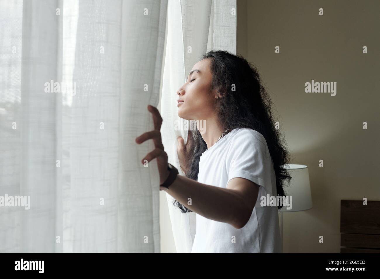 Calm handsome young man with long curly hair opening curtains and enjoying first rays of sun Stock Photo
