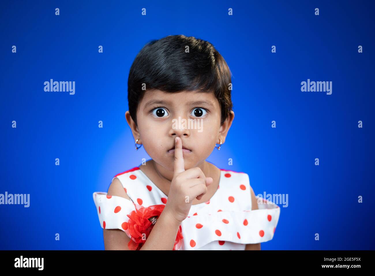 Cute girl kid saying keep silence quite by placing finger on mouth on blue studio background by looking at camera. Stock Photo
