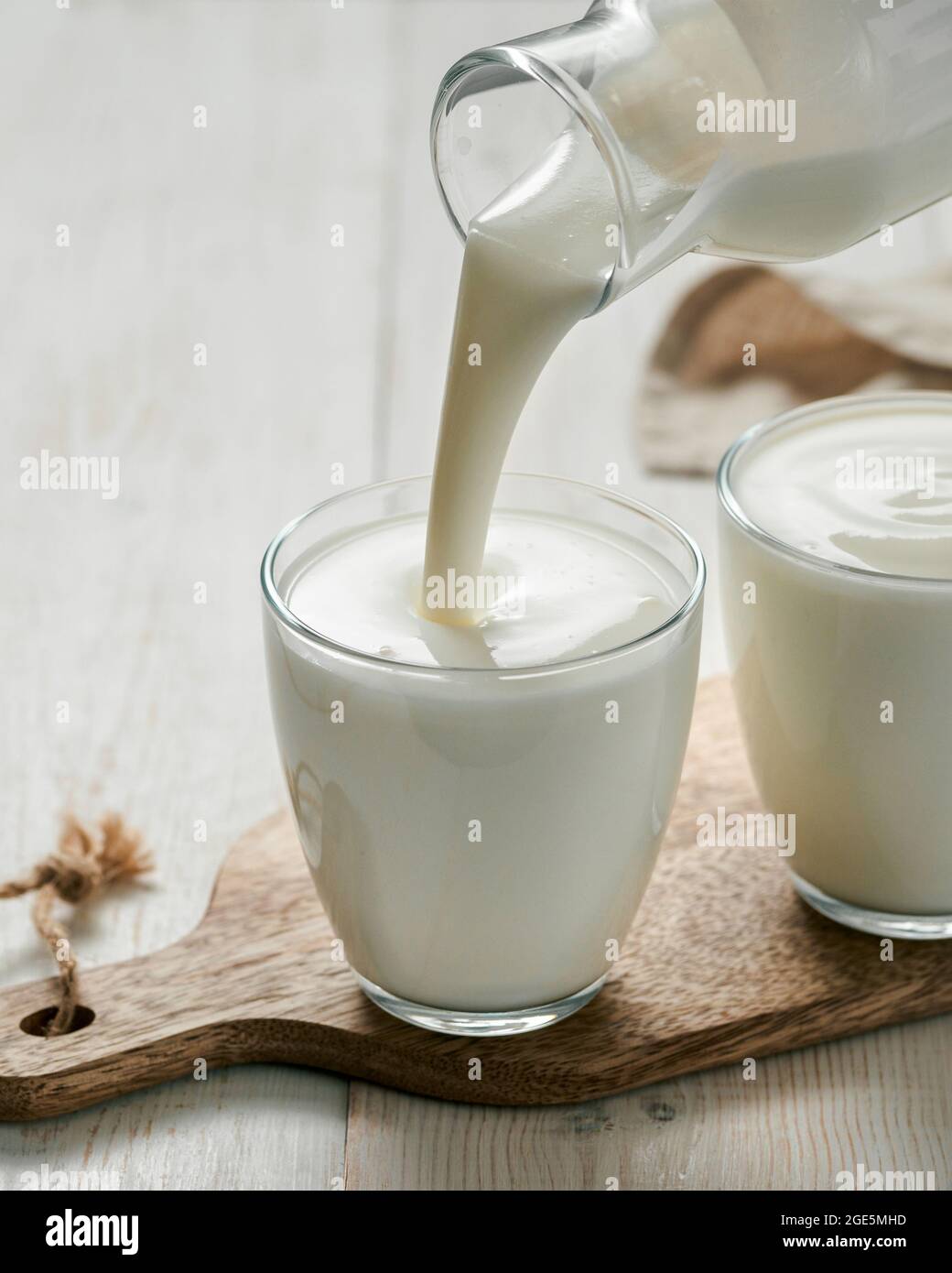 Pouring kefir, buttermilk or yogurt with probiotics. Yogurt flowing from glass bottle on white wooden background. Probiotic cold fermented dairy milk drink. Vertical Stock Photo