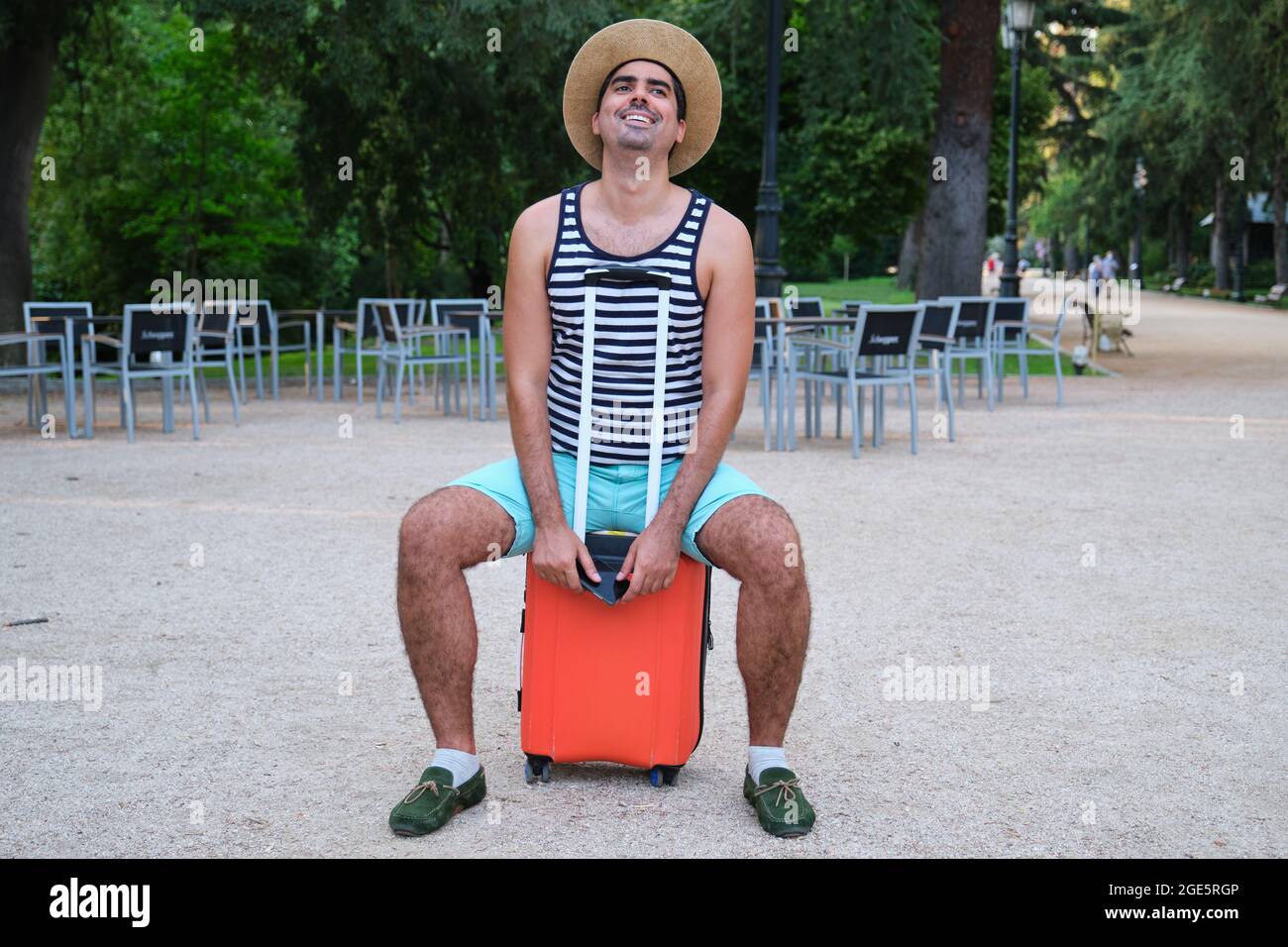 Tourist man smiling sitting on suitcase in a park. Stock Photo