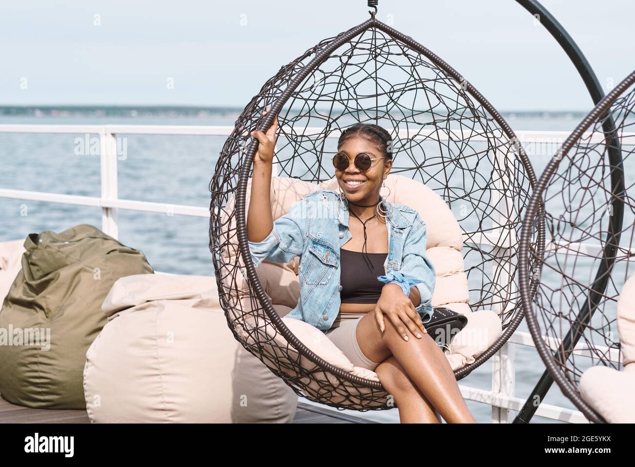 Portrait of attractive smiling young woman in sunglasses relaxing in hanging chair outdoors at party Stock Photo