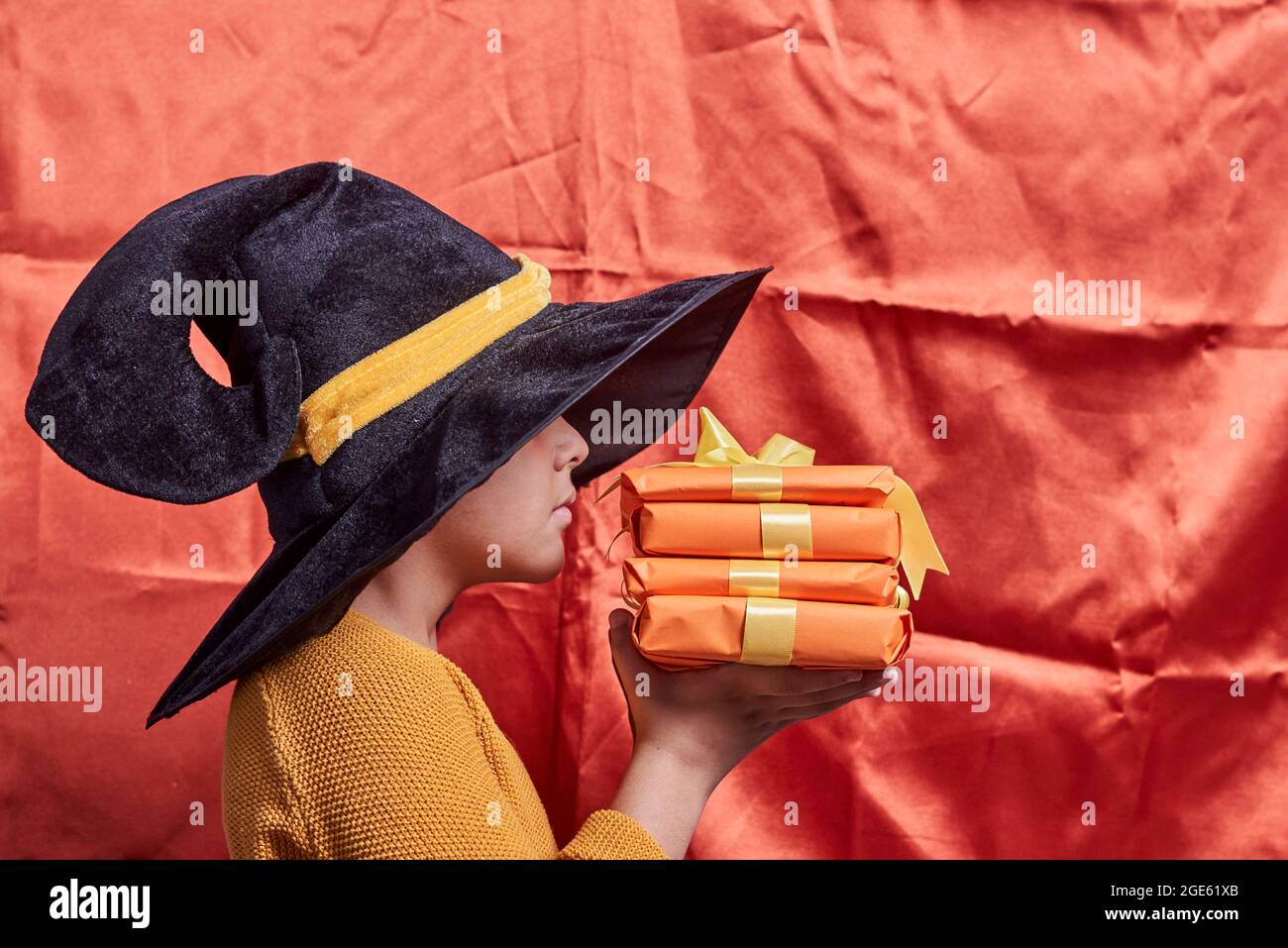 A little kid in witch cap holding a pile of gift boxes. Halloween gifts in hands of a boy over orange background Stock Photo
