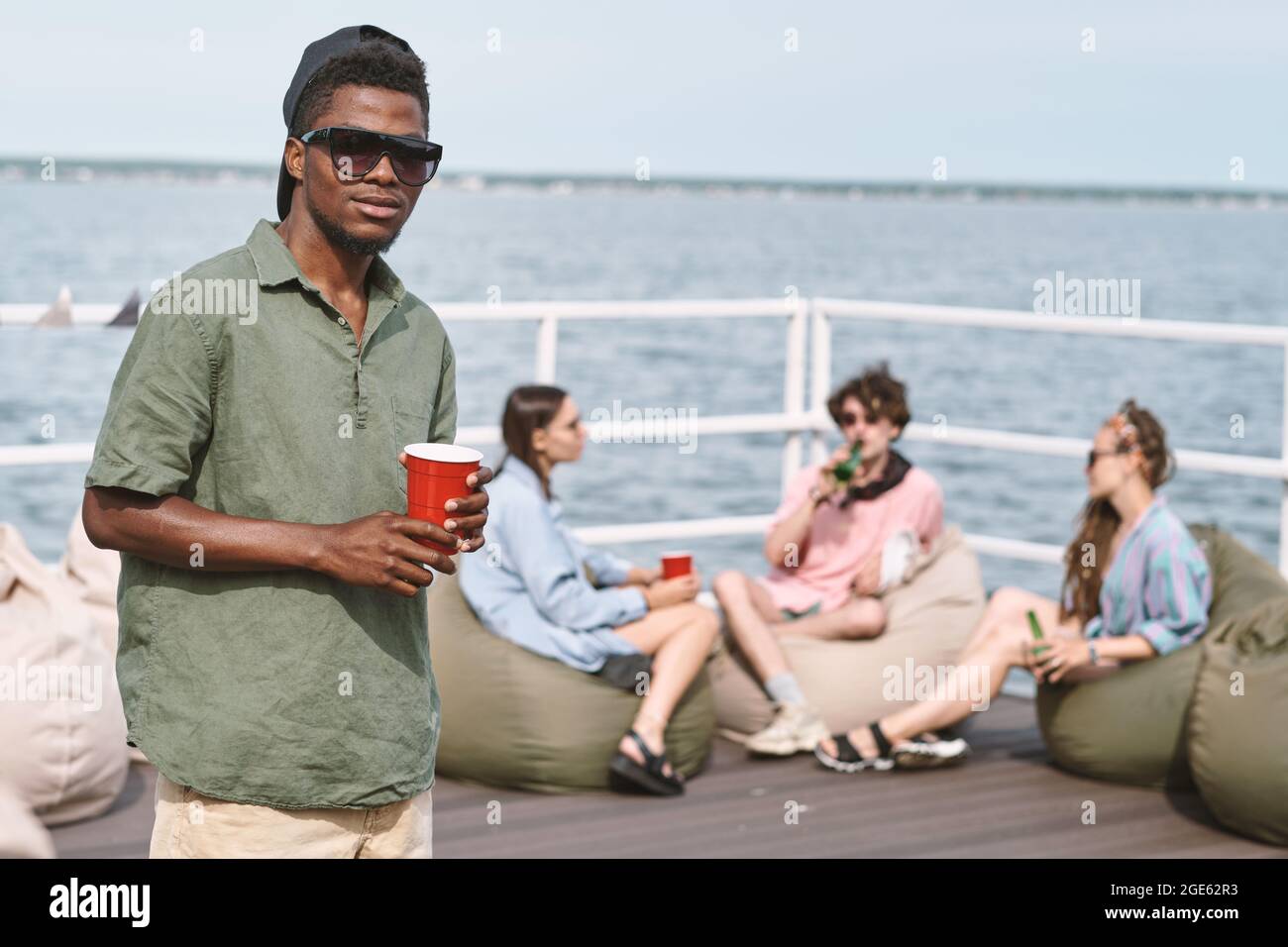 Young man in cap and sunglasses holding red plastic cup, his friend relaxing on bean bags in background Stock Photo