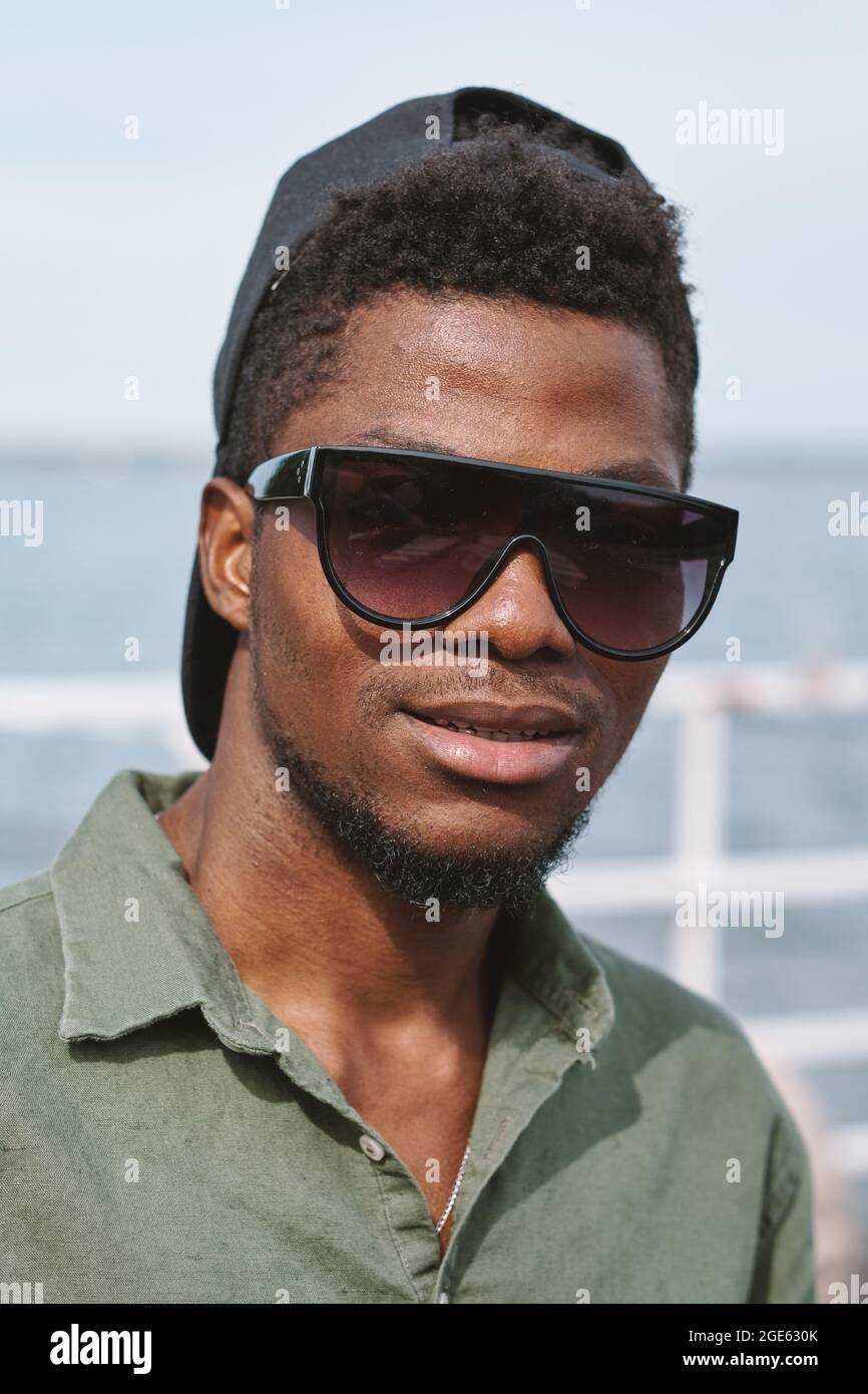 Face of handsome serious young man in sunglasses and cap looking at camera Stock Photo