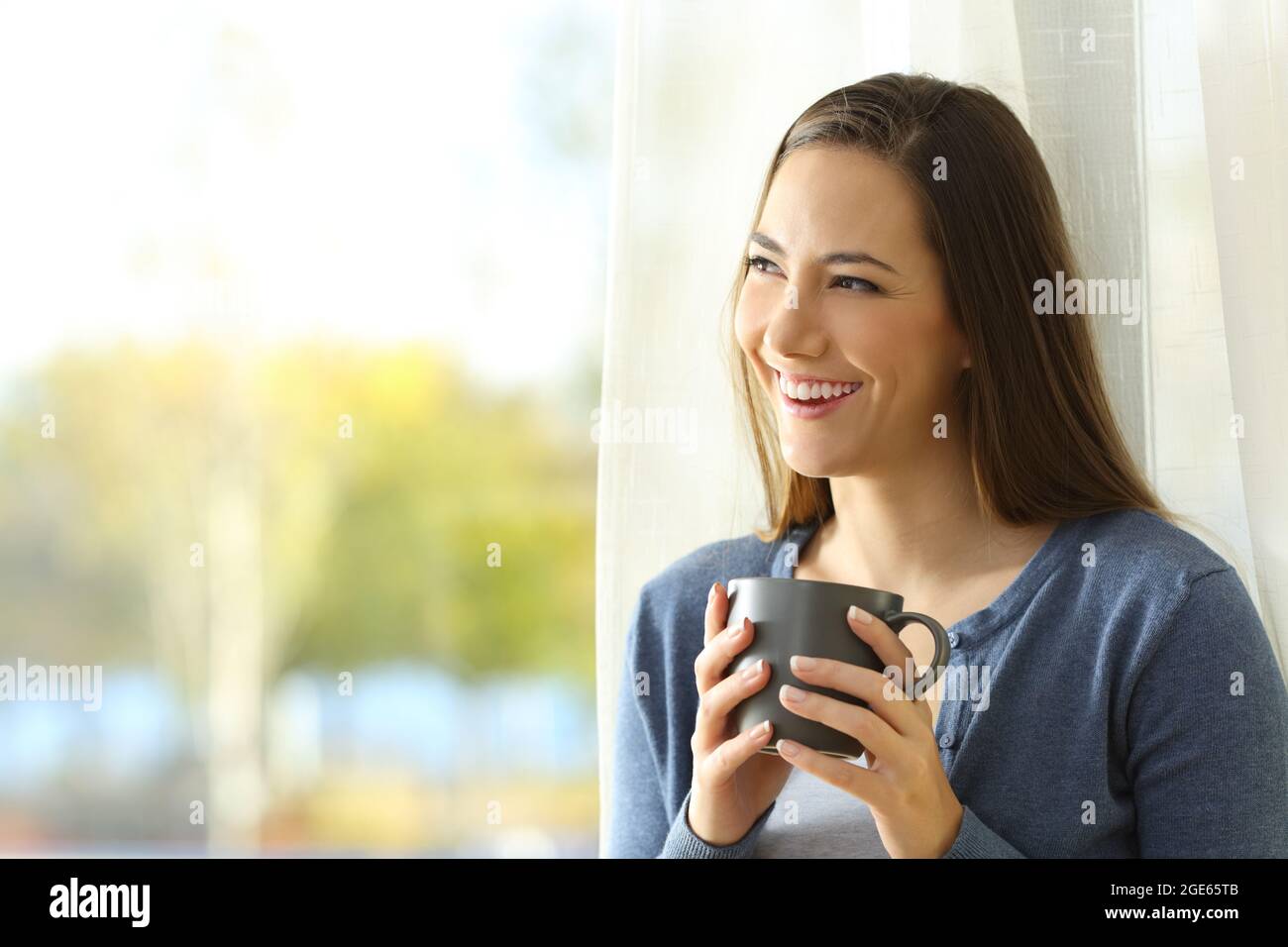 Happy pensive woman looks at side beside a window holding a coffee cup at home Stock Photo