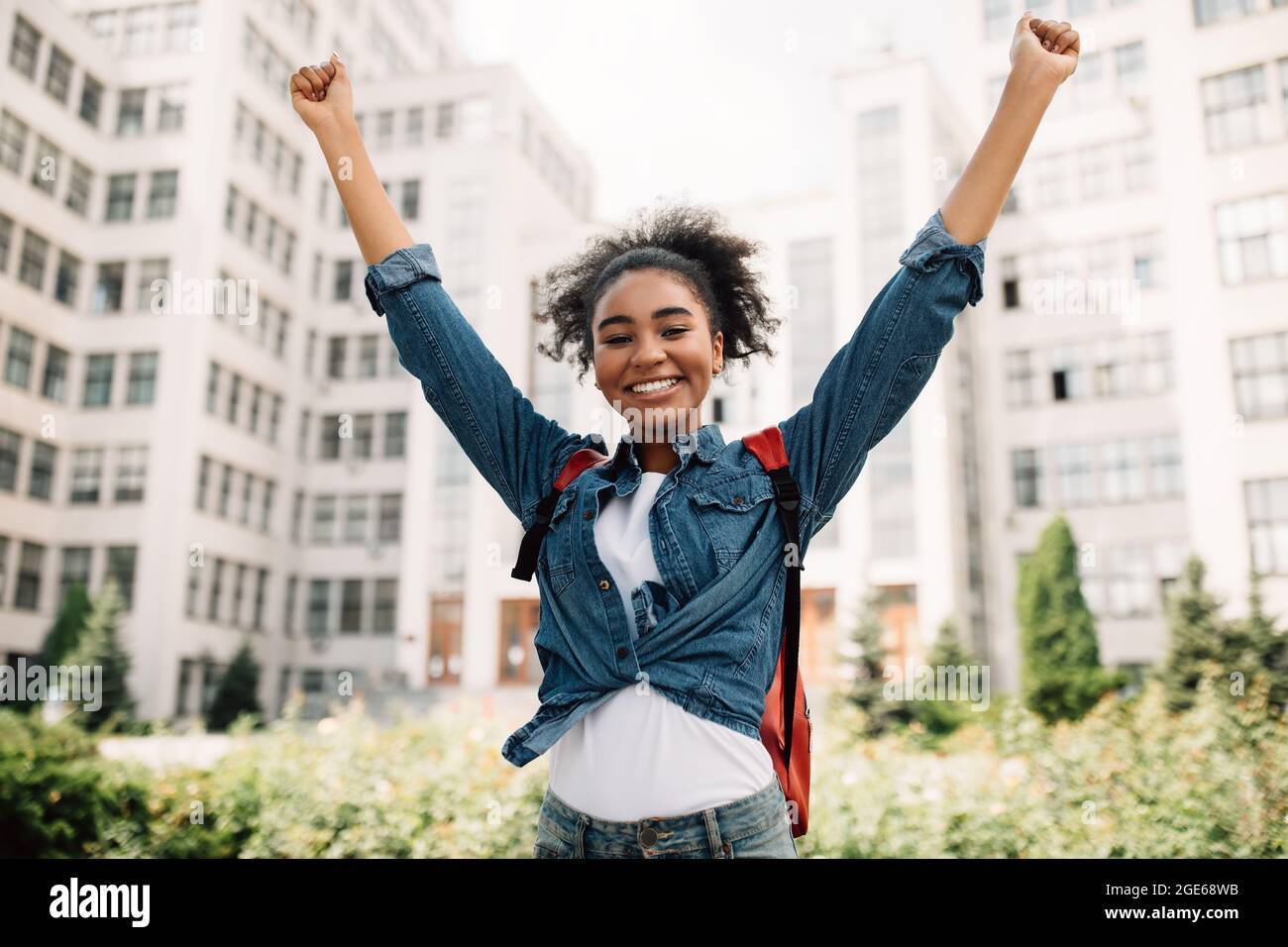 Passed Exam. Joyful Black Student Girl Shaking Fists Raising Arms Celebrating First Day In College Posing With Backpack Near University Building Outdo Stock Photo