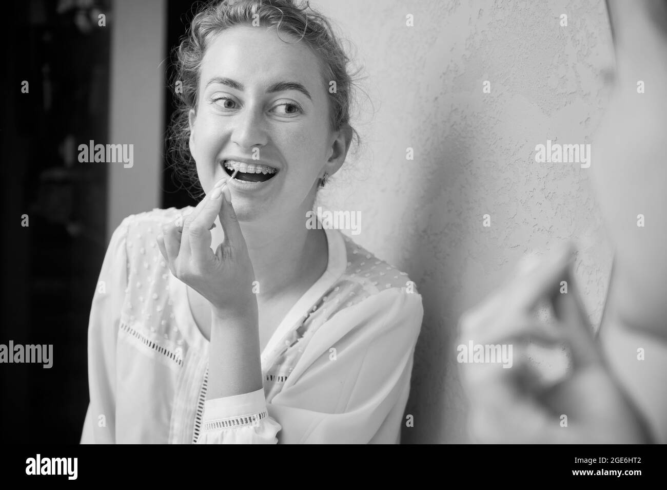 Portrait of a beautiful woman in dentist's office demonstrating interdental brushing, looking in huge mirror, wearing dental ceramic and metal brackets. Black and white image Stock Photo