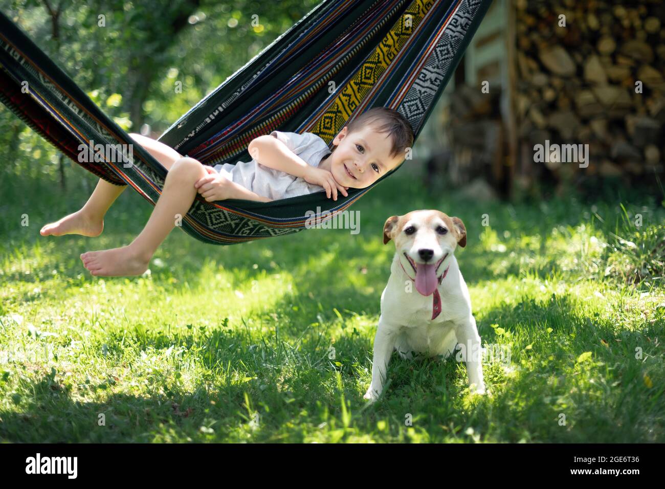 Small kid on hammock with white dog puppy breed jack russel terrier on summer backyard. Happy childhood concept Stock Photo