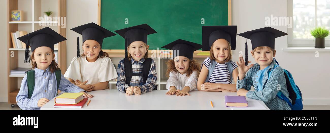 Banner with group of happy school kids in academic caps standing by table in classroom Stock Photo
