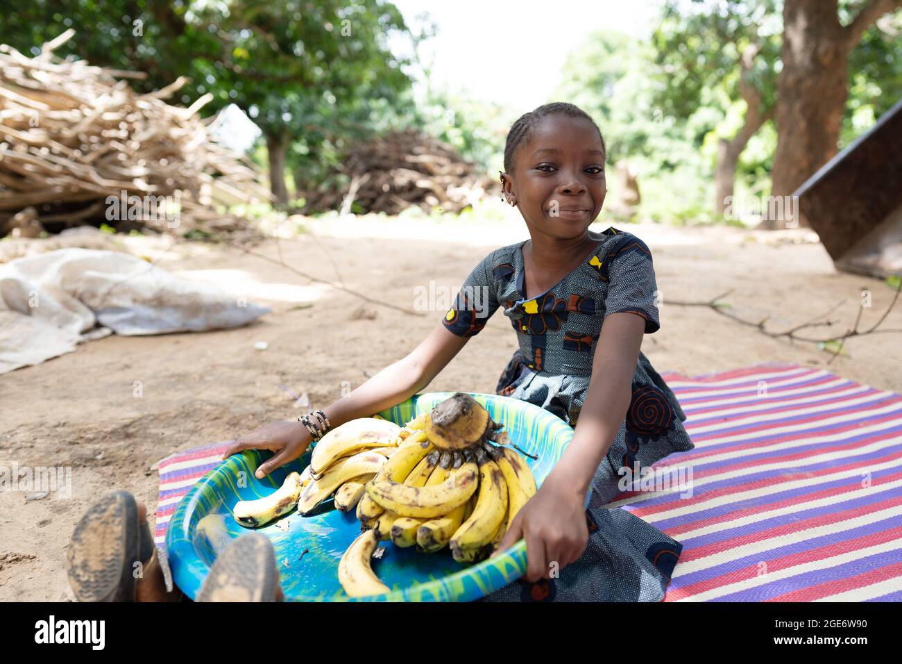 Pretty little African girl holding a big plate full of ripe bananas on her knees, smirking at camera Stock Photo