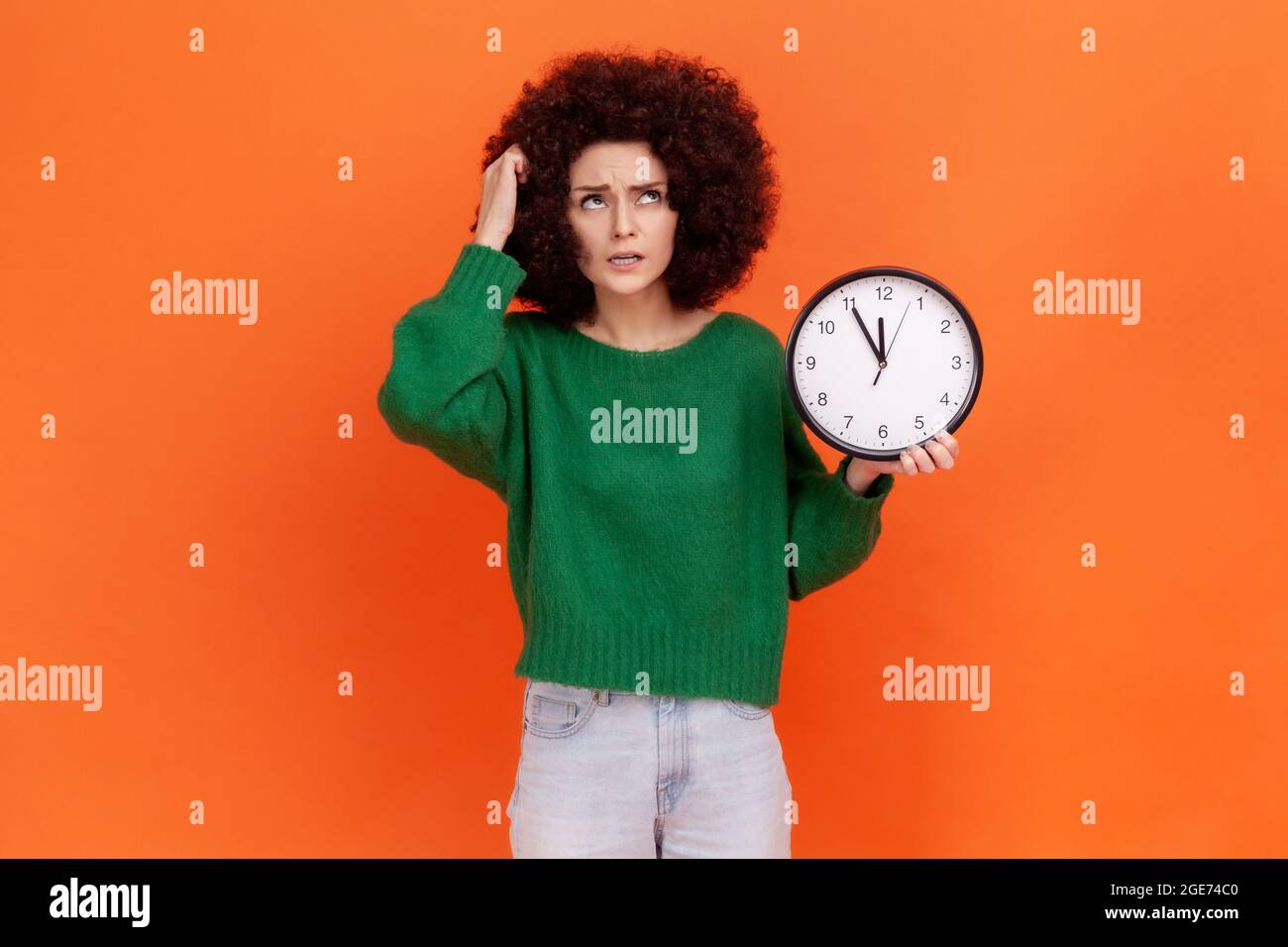 Portrait of pensive woman with curly hair wearing green casual style sweater holding wall clock and scratching her head, deadline, punctuality. Indoor Stock Photo