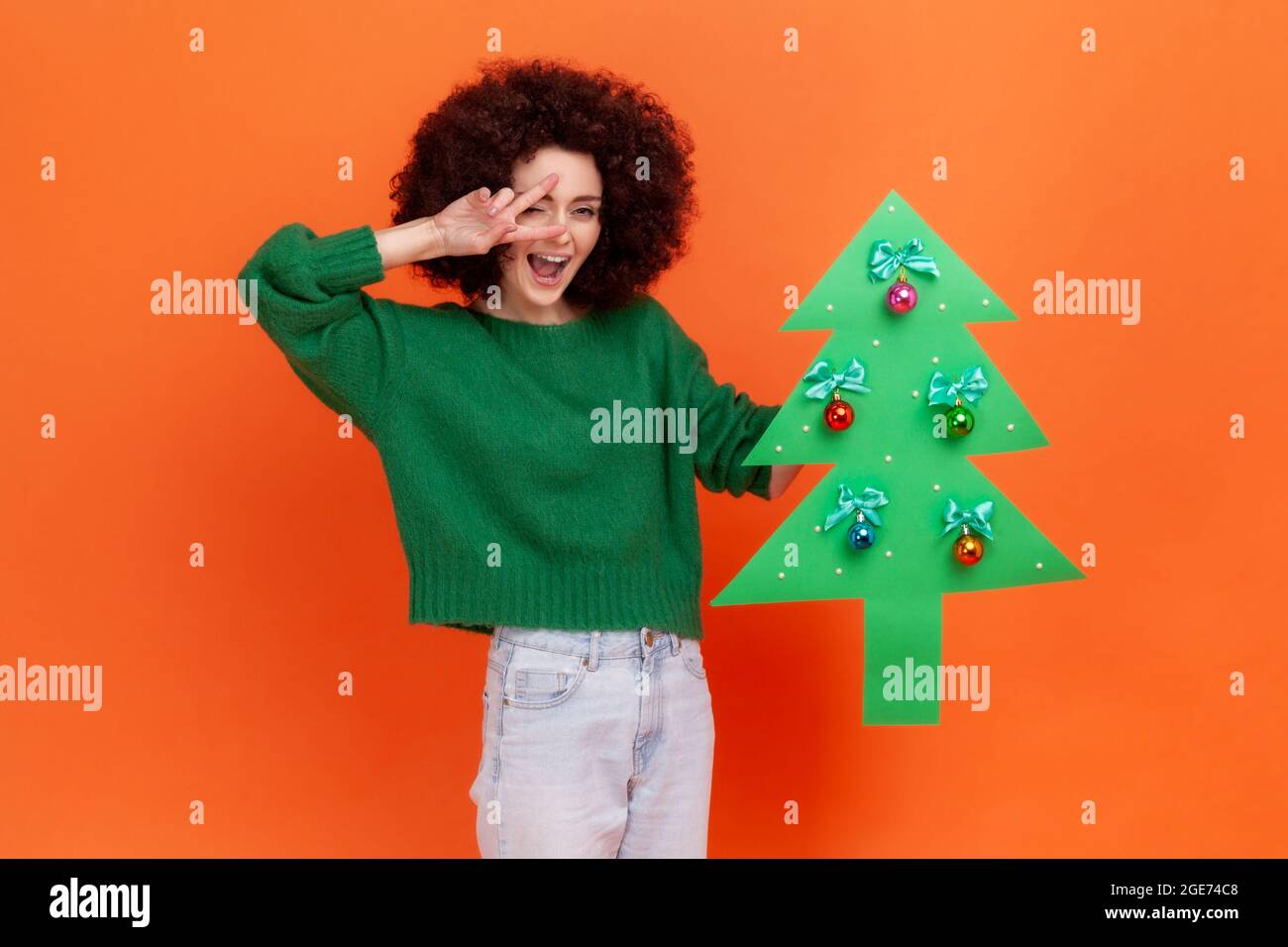Portrait of positive woman with Afro hairstyle wearing green casual style sweater holding paper christmas tree and showing v sign, happy expression. I Stock Photo