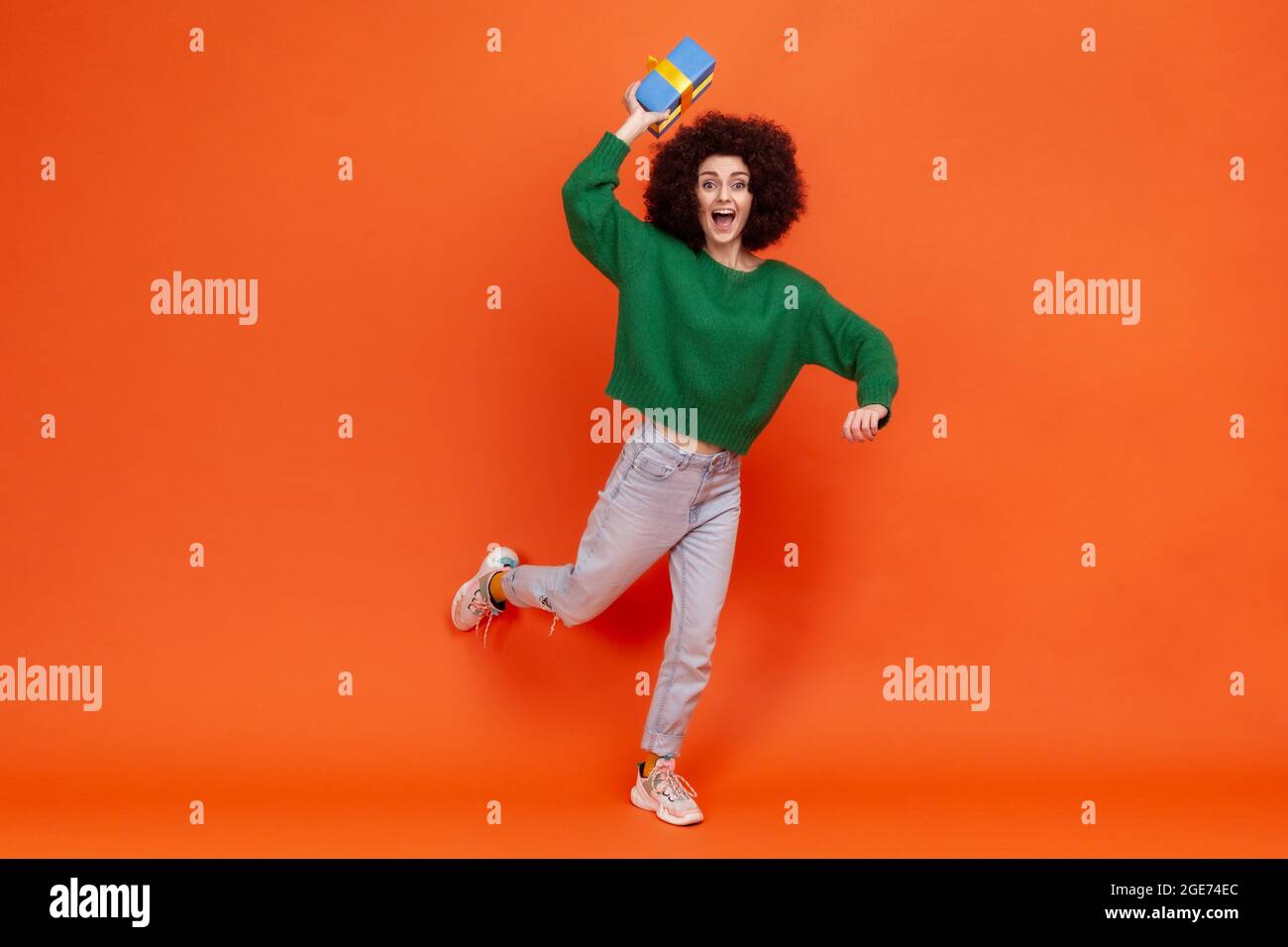 Full length portrait of happy woman with curly hair wearing green casual style sweater throwing blue wrapped present box, has excited expression. Indo Stock Photo