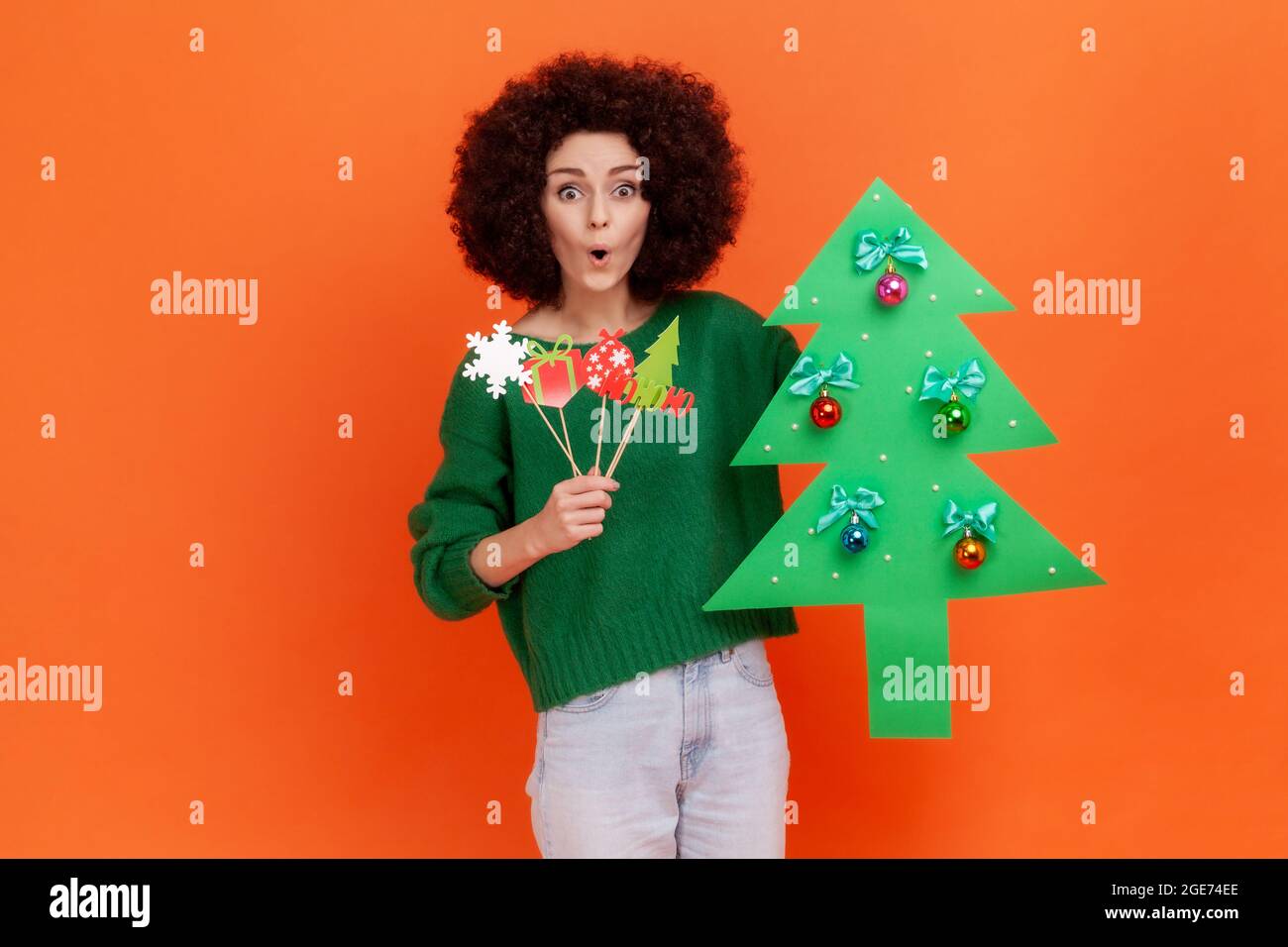 Amazed woman with curly hair wearing green casual style sweater holding mask on sticks and Christmas tree, having shocked expression. Indoor studio sh Stock Photo